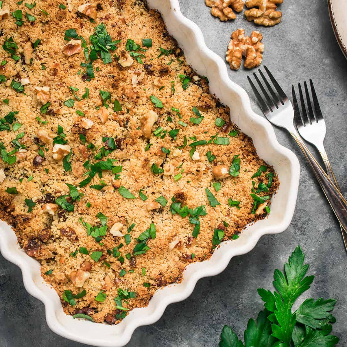 A baked Butternut Squash Casserole topped with golden breadcrumbs, chopped parsley, and walnut pieces sits in a scalloped white dish. Two forks, some walnuts, and parsley leaves are placed nearby on a gray surface.