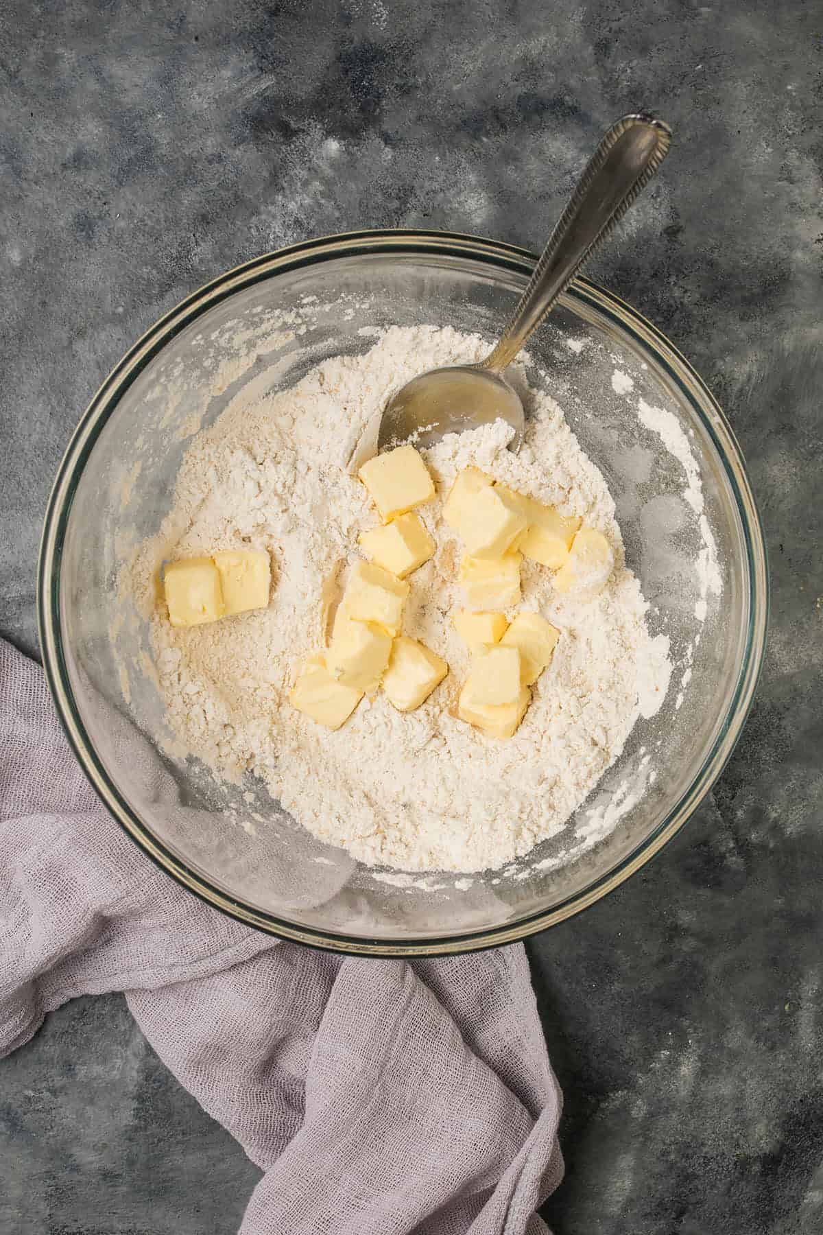 A glass bowl with flour and cubed butter, partially mixed with a metal spoon—perfect for starting a Butternut Squash Casserole. A light purple cloth rests beside the bowl on a dark textured surface.