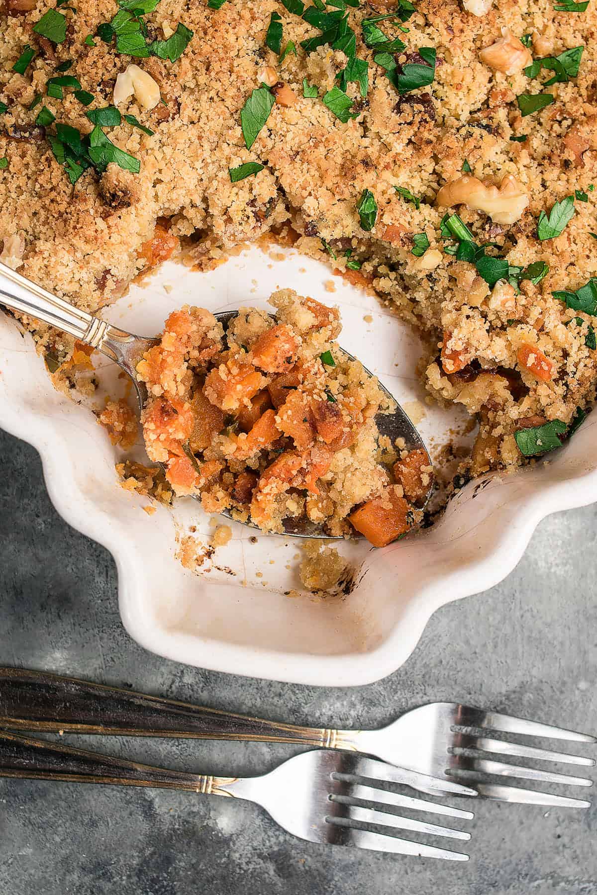 A ceramic baking dish holds a golden-brown Butternut Squash Casserole crumble topped with chopped parsley. A serving spoon reveals diced vegetables inside, while three metal forks rest nearby on a gray surface.