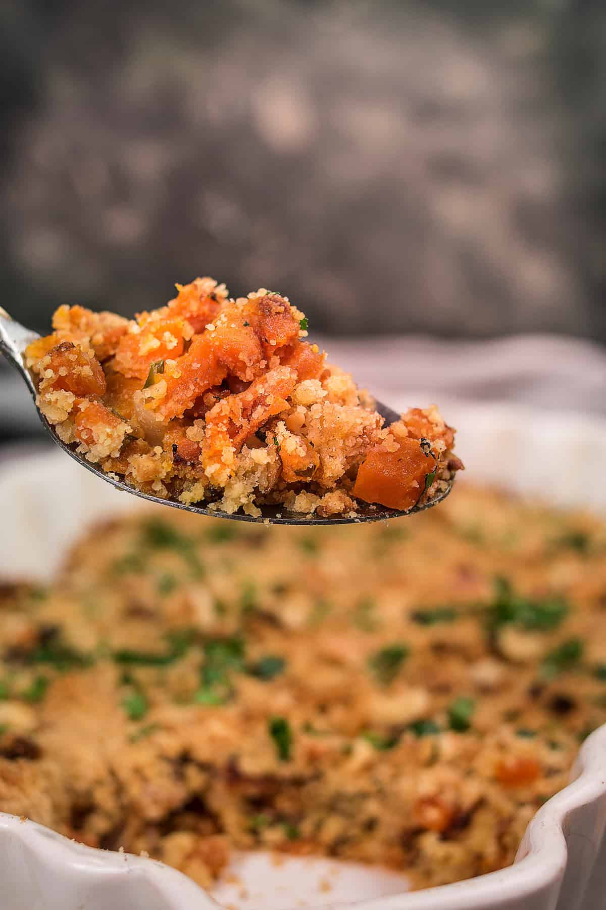 A close-up of a spoonful of Butternut Squash Casserole topped with golden breadcrumbs, lifted above a baking dish filled with more casserole and garnished with chopped herbs.