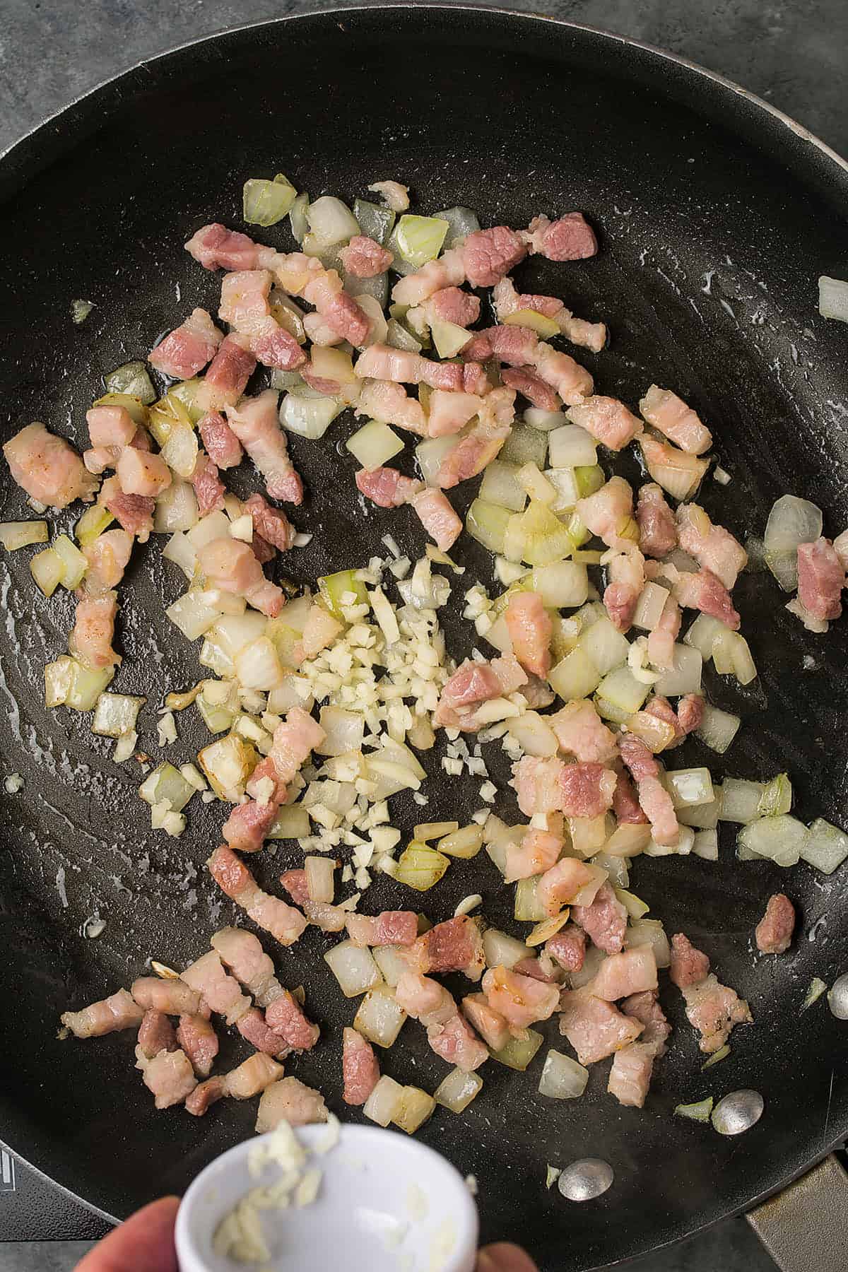 Chopped bacon and diced onions cooking in a skillet, with a hand adding minced garlic from a small white bowl—perfect aromatics for starting a delicious Butternut Squash Casserole, all sizzling together on the stovetop.