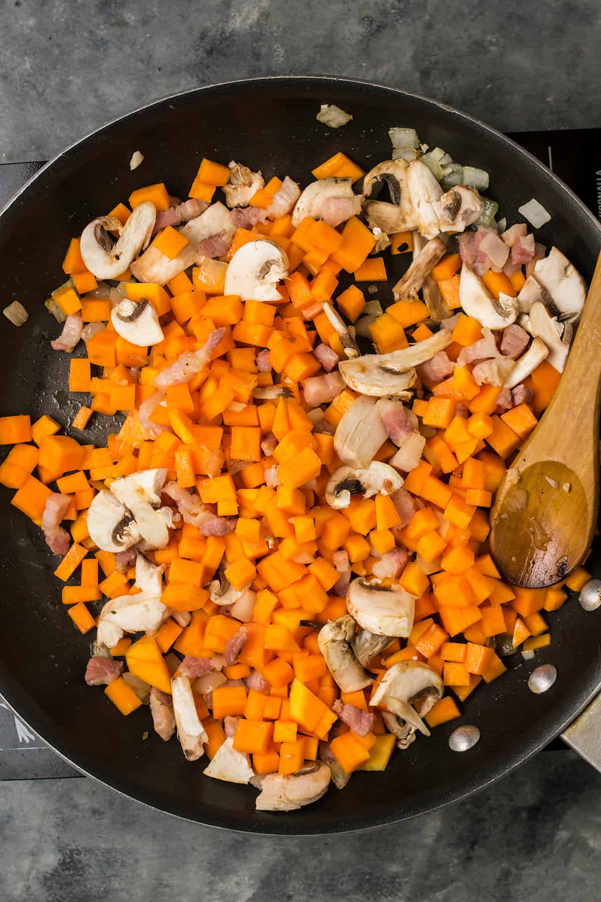 A frying pan filled with chopped carrots, mushrooms, onions, and pieces of bacon being sautéed for a savory Butternut Squash Casserole, with a wooden spoon resting on the side.