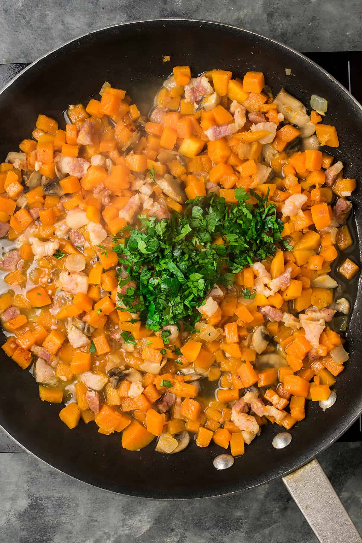 A skillet filled with sautéed diced butternut squash, bacon, and mushrooms, reminiscent of a hearty Butternut Squash Casserole, topped with a mound of freshly chopped parsley in the center.