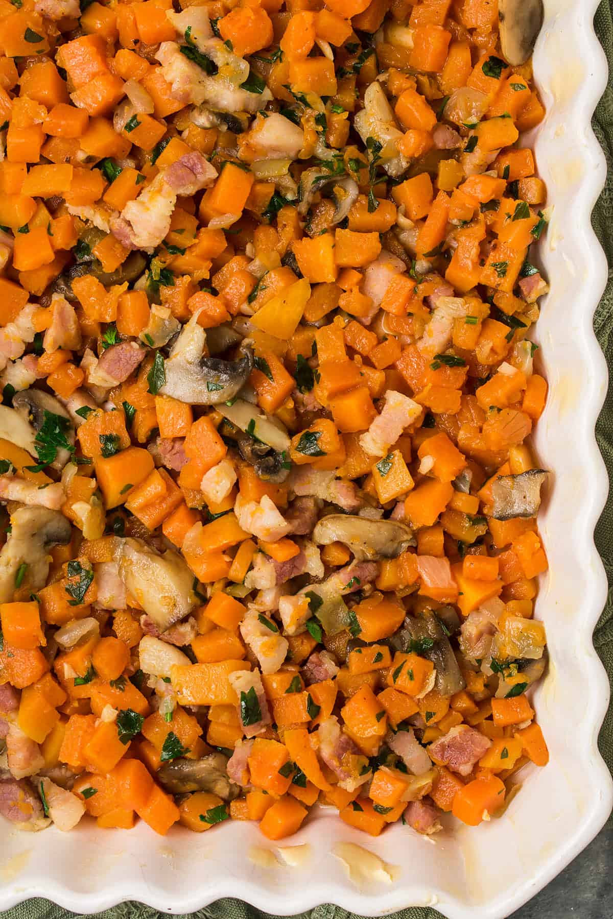 A close-up of a Butternut Squash Casserole filled with diced orange vegetables, mushrooms, bacon pieces, and fresh herbs, all mixed together in a white scalloped-edge baking dish.