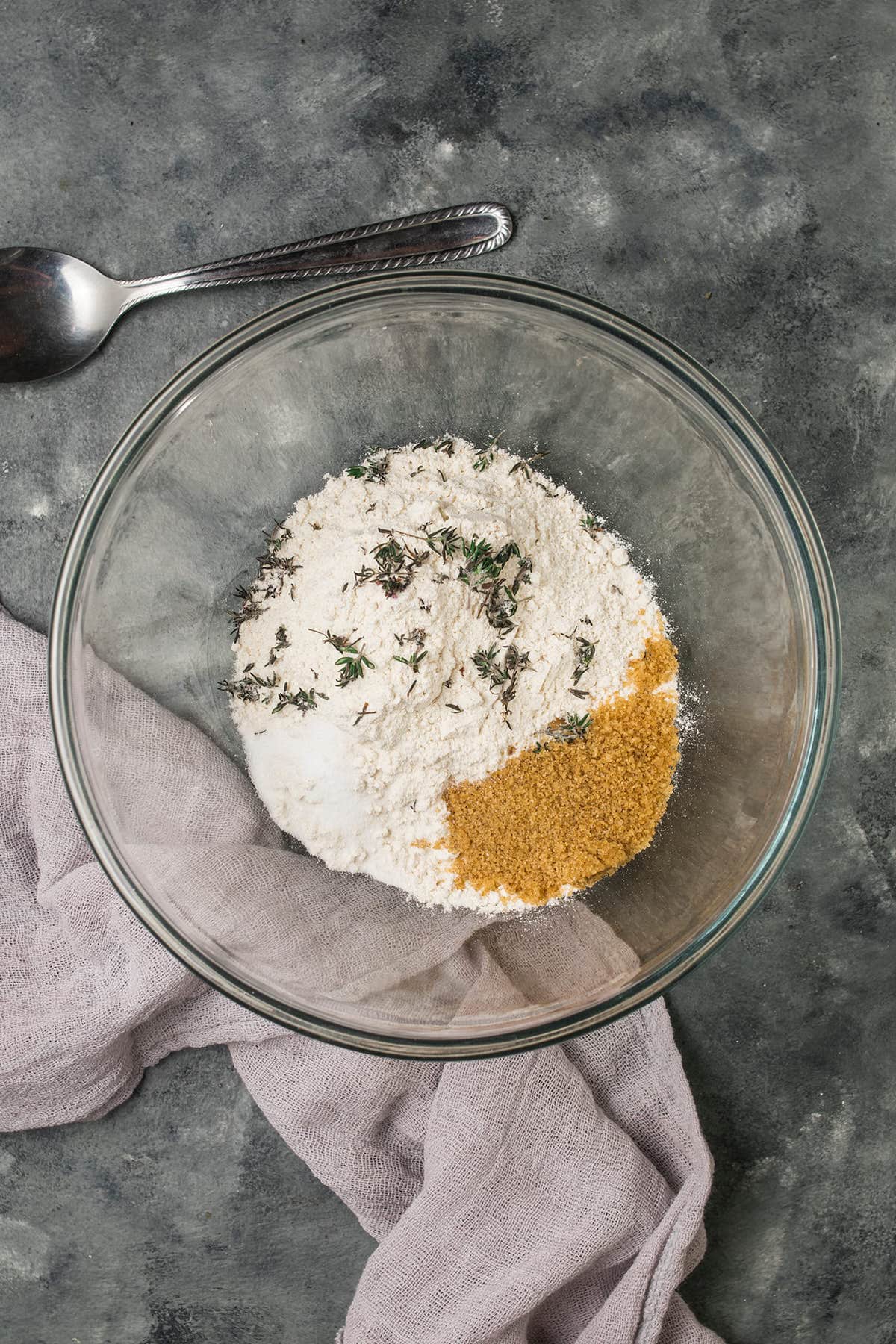 A glass bowl with flour, brown sugar, and fresh thyme sits on a gray surface, ready for your Butternut Squash Casserole—alongside a silver spoon and a light purple cloth.