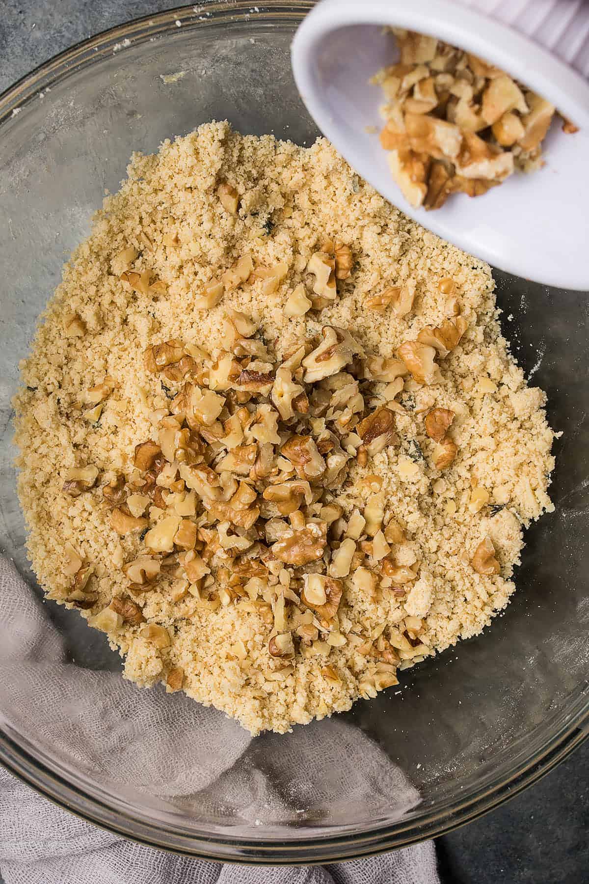 A glass bowl filled with a crumbly flour mixture for Butternut Squash Casserole, with chopped walnuts being poured in from a small white ramekin, and a beige cloth partly visible beside the bowl.