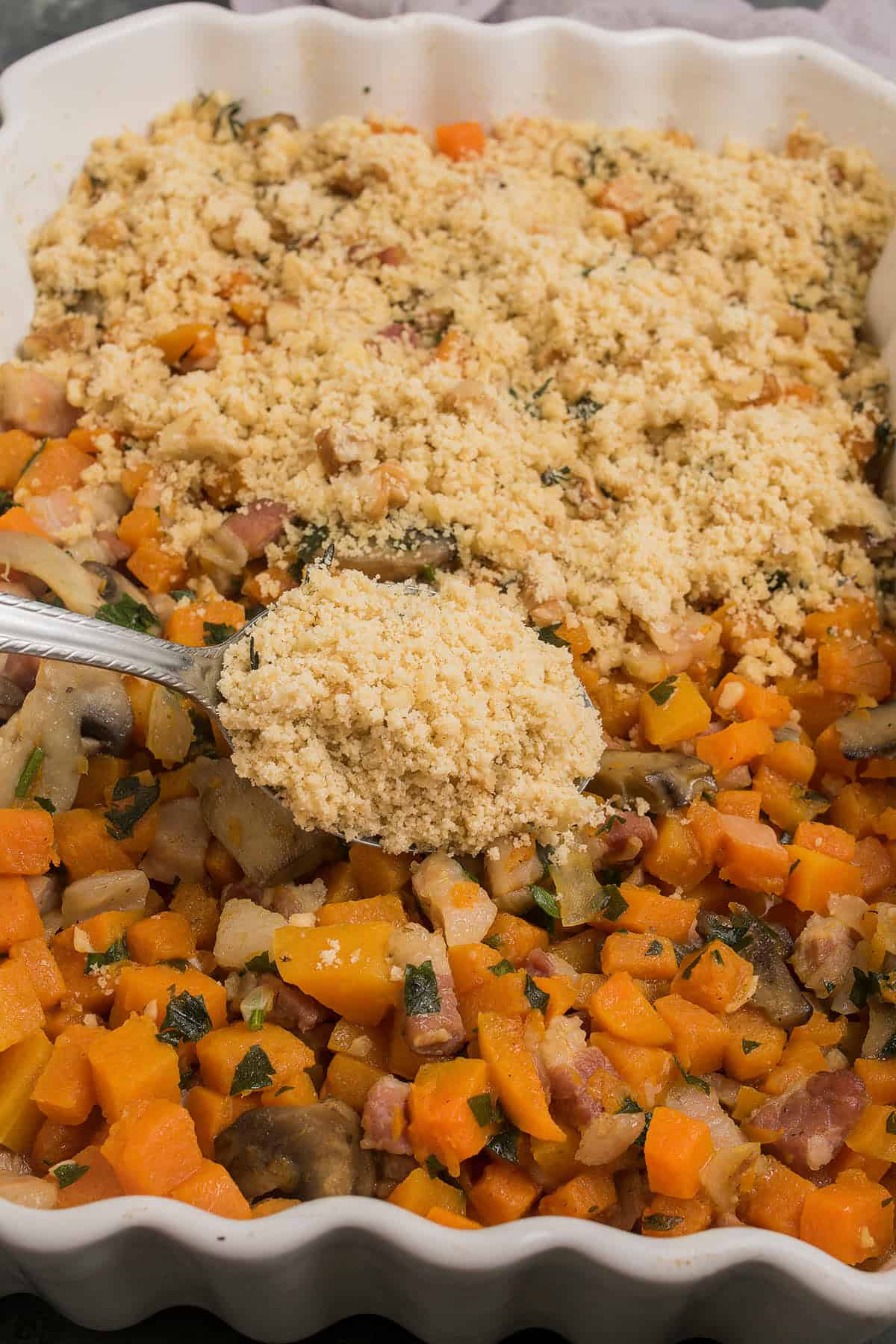 A close-up of a Butternut Squash Casserole topped with a golden breadcrumb mixture in a white baking dish. A spoon scoops up the crumb topping, revealing tender, diced vegetables underneath.