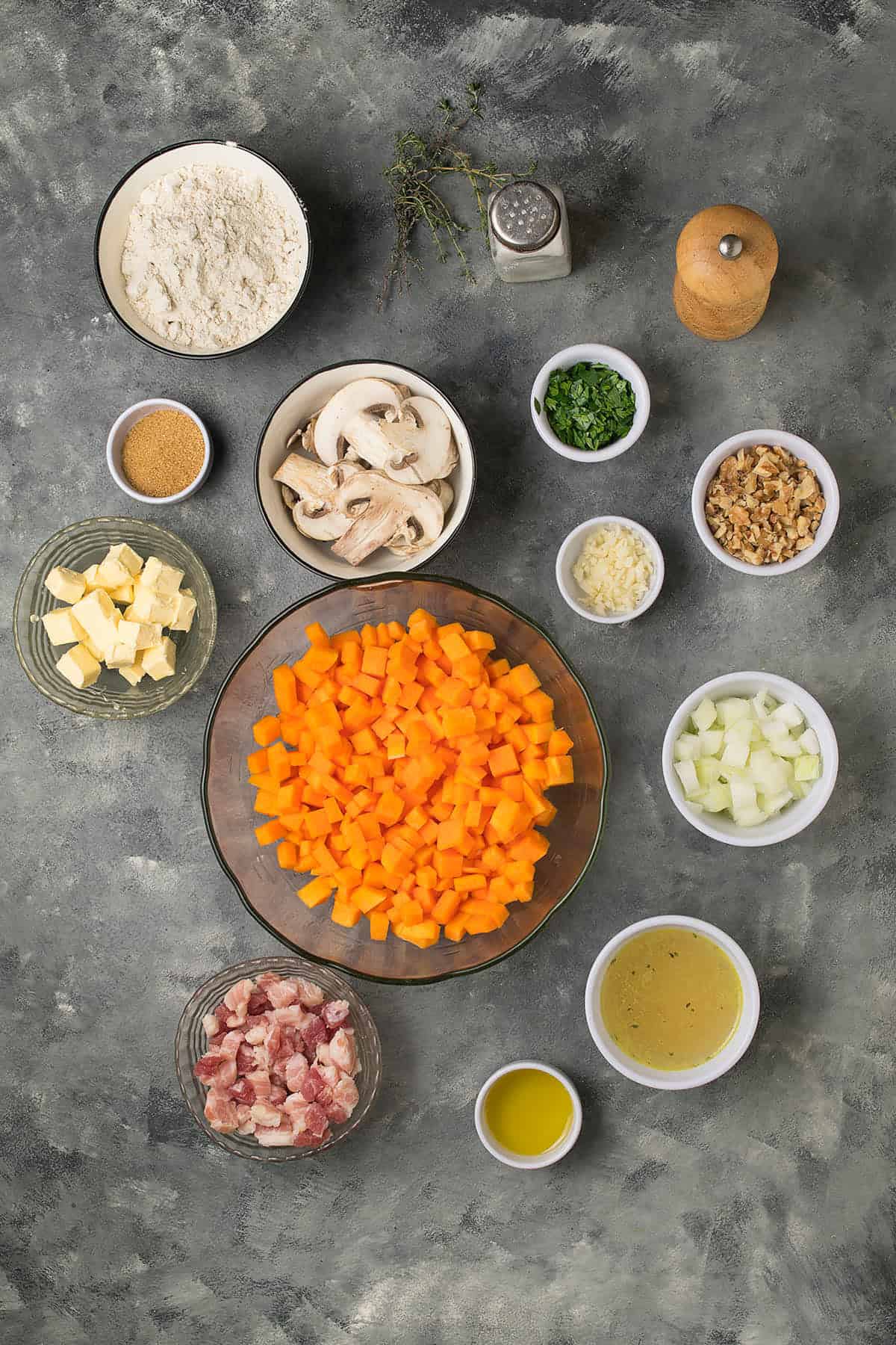 A flat lay of various ingredients in bowls on a gray surface, perfect for making Butternut Squash Casserole, including chopped carrots, onions, mushrooms, butter, flour, broth, parsley, cubed meat, garlic, seasonings, and olive oil.