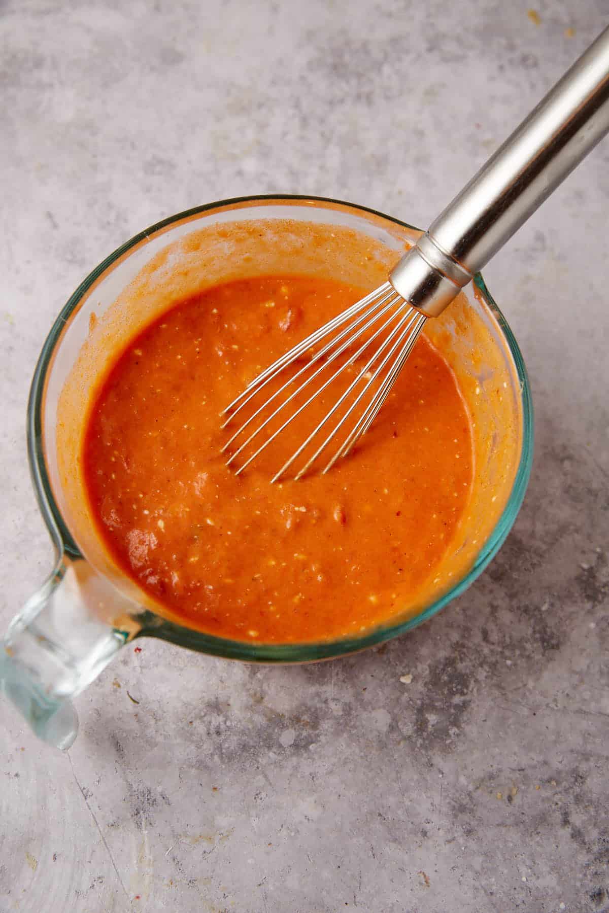 A glass measuring cup filled with orange-red sauce for Baked Ravioli is being whisked with a metal whisk, placed on a light gray textured surface.