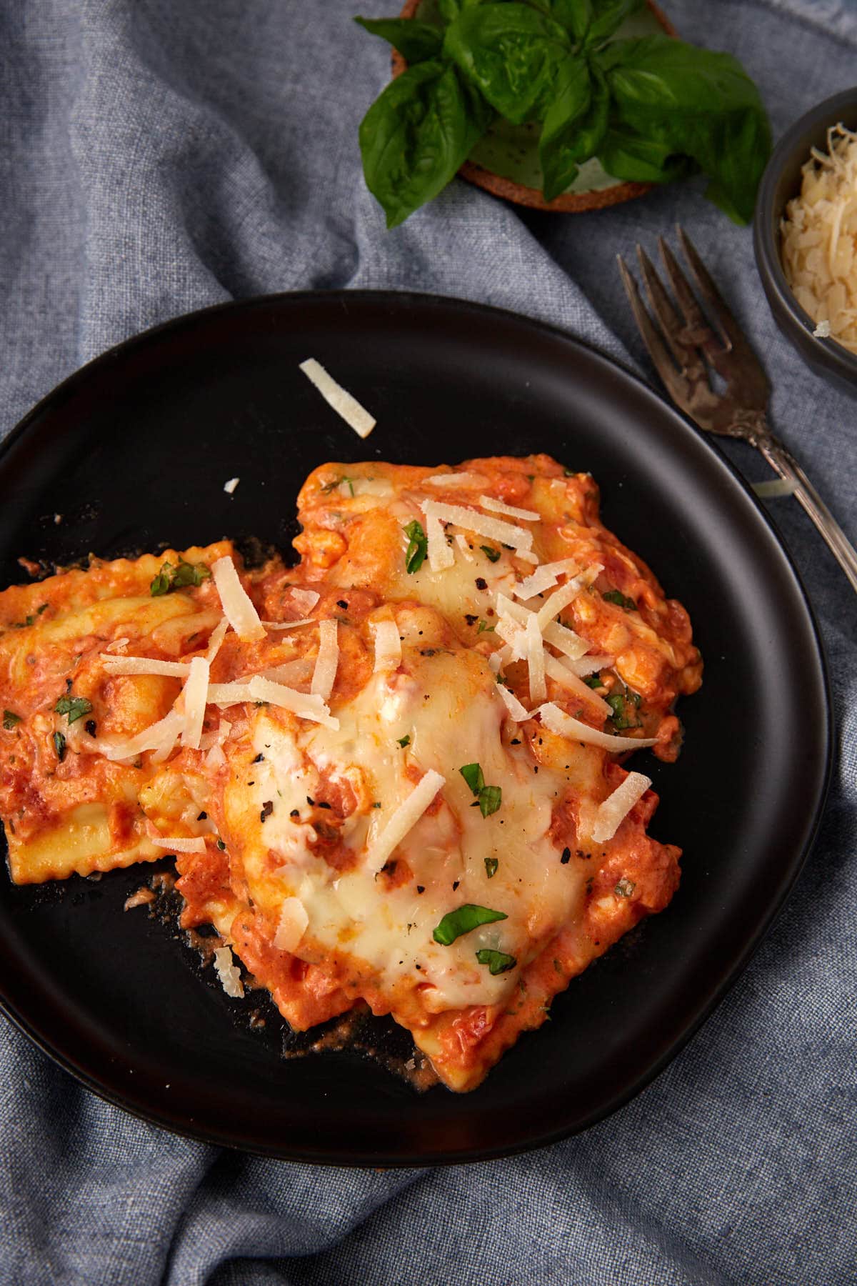 A black plate with cheesy baked ravioli topped with tomato sauce, shredded Parmesan, and fresh basil. A fork, grated cheese, and basil leaves sit nearby on a gray cloth background for the perfect baked ravioli experience.