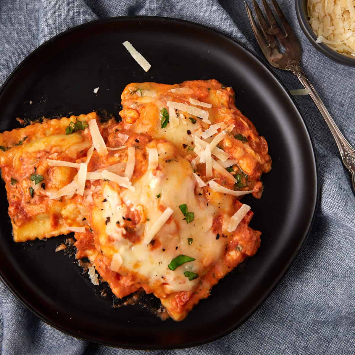 Three pieces of baked ravioli covered in melted cheese and tomato sauce sit on a black plate, topped with shredded parmesan and herbs. A fork and a small bowl of grated cheese are nearby on a blue cloth, highlighting the perfect baked ravioli dish.