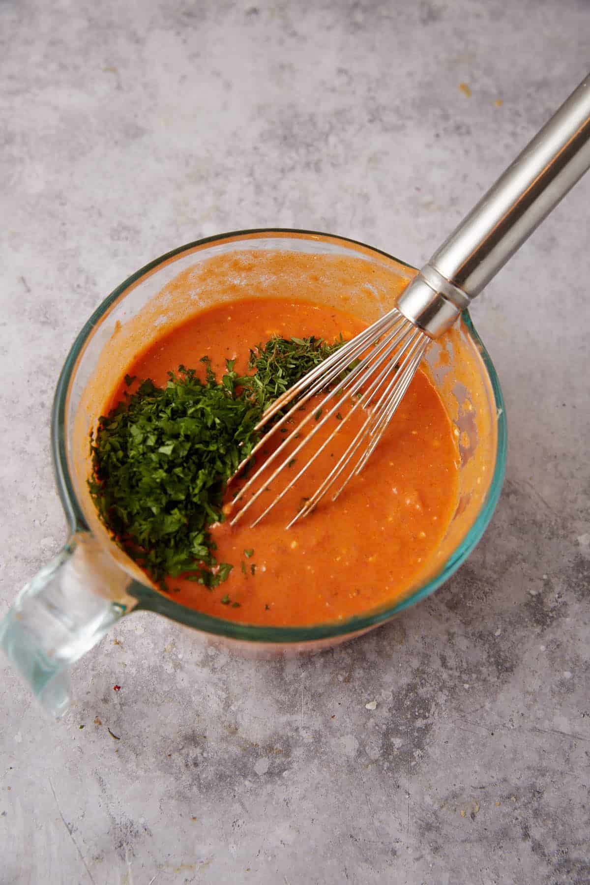 A glass measuring cup with an orange sauce mixture for baked ravioli, freshly chopped green herbs, and a metal whisk on a light gray textured surface.