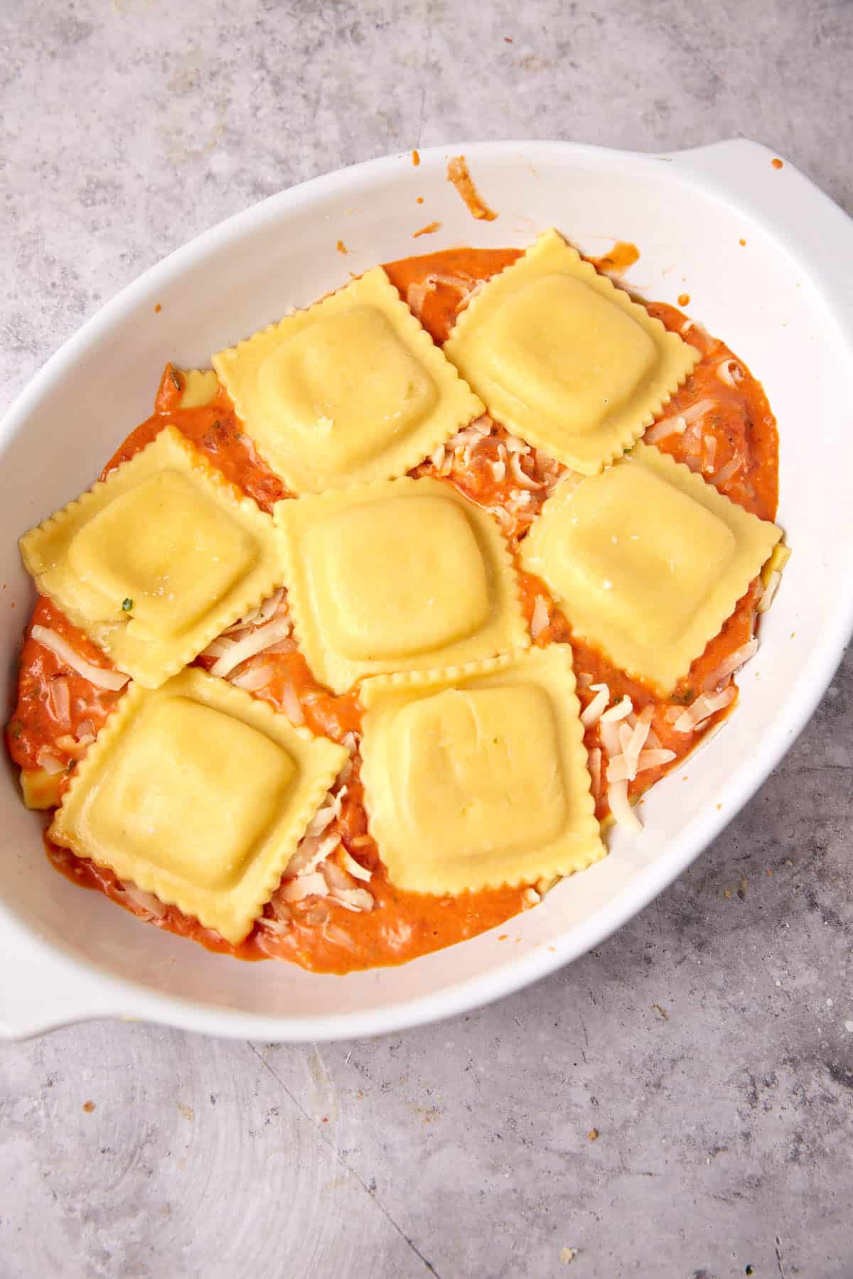 A white oval baking dish with seven uncooked ravioli for baked ravioli, resting on shredded cheese and tomato sauce, set on a gray textured surface.