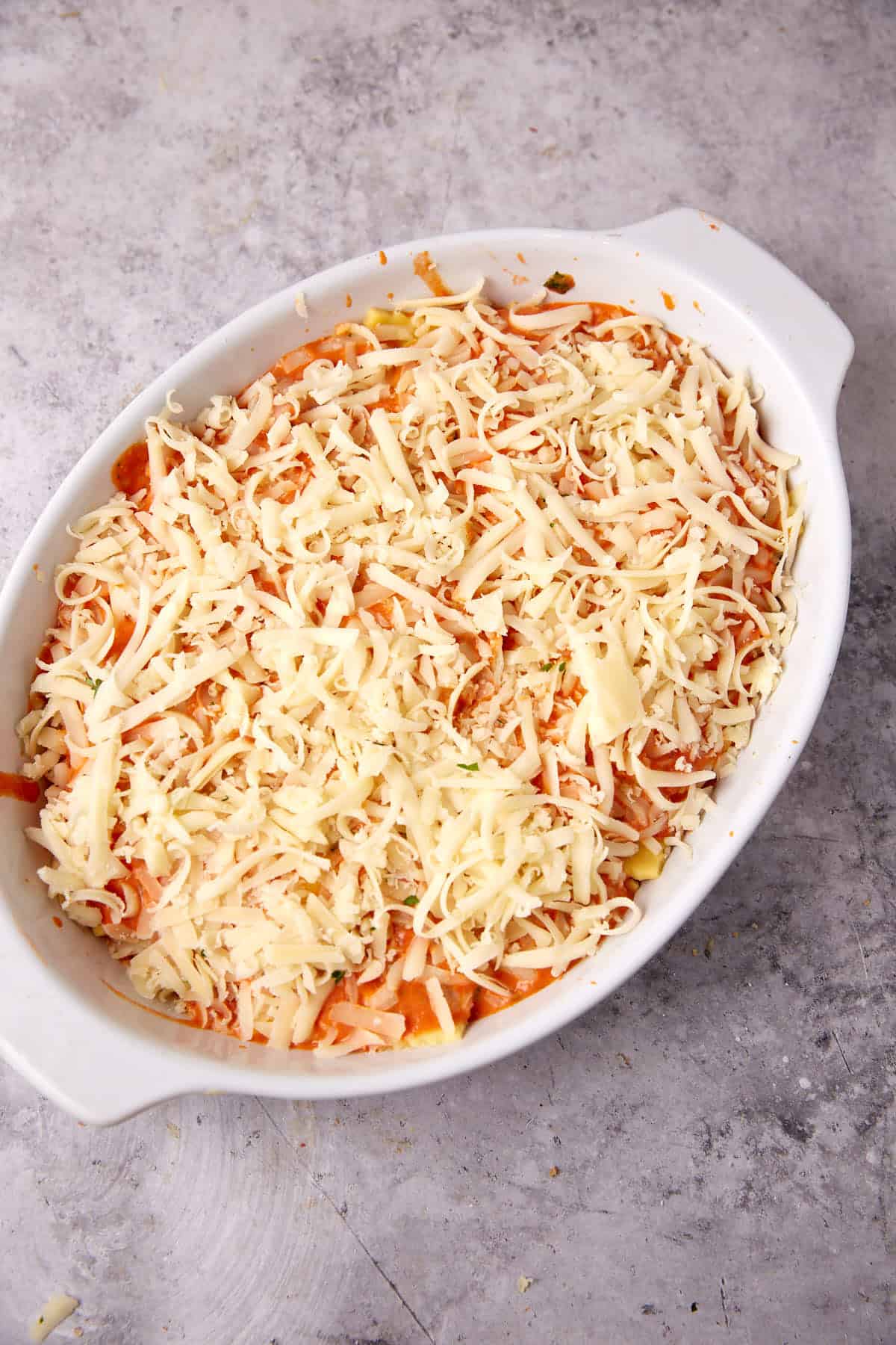 An oval white baking dish filled with baked ravioli casserole, generously topped with shredded cheese, set on a light gray textured surface.