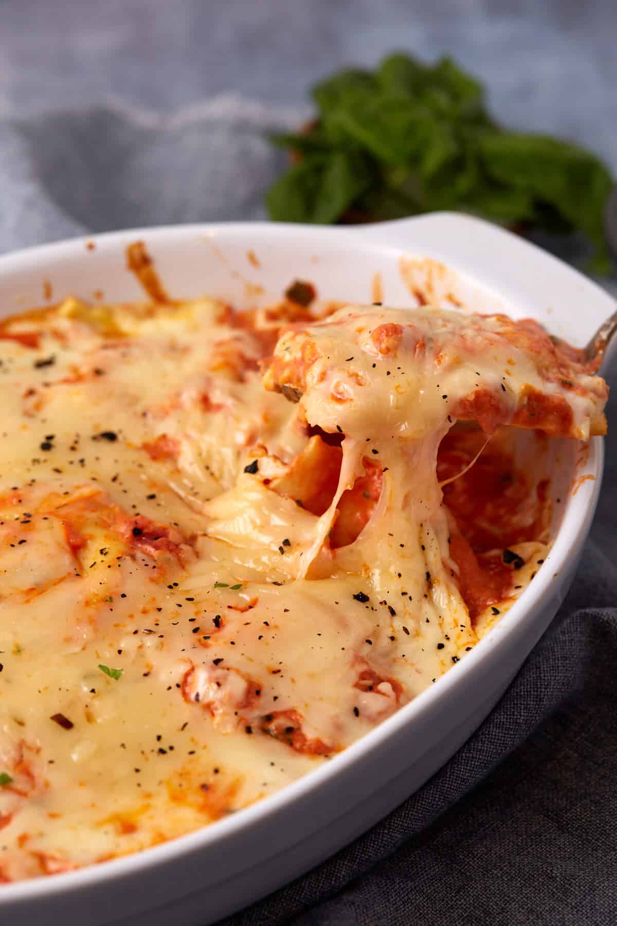 A close-up of baked ravioli lasagna in a white dish, with melted cheese being lifted by a serving utensil, showing stretchy cheese and layers of sauce beneath. Fresh herbs are blurred in the background.
