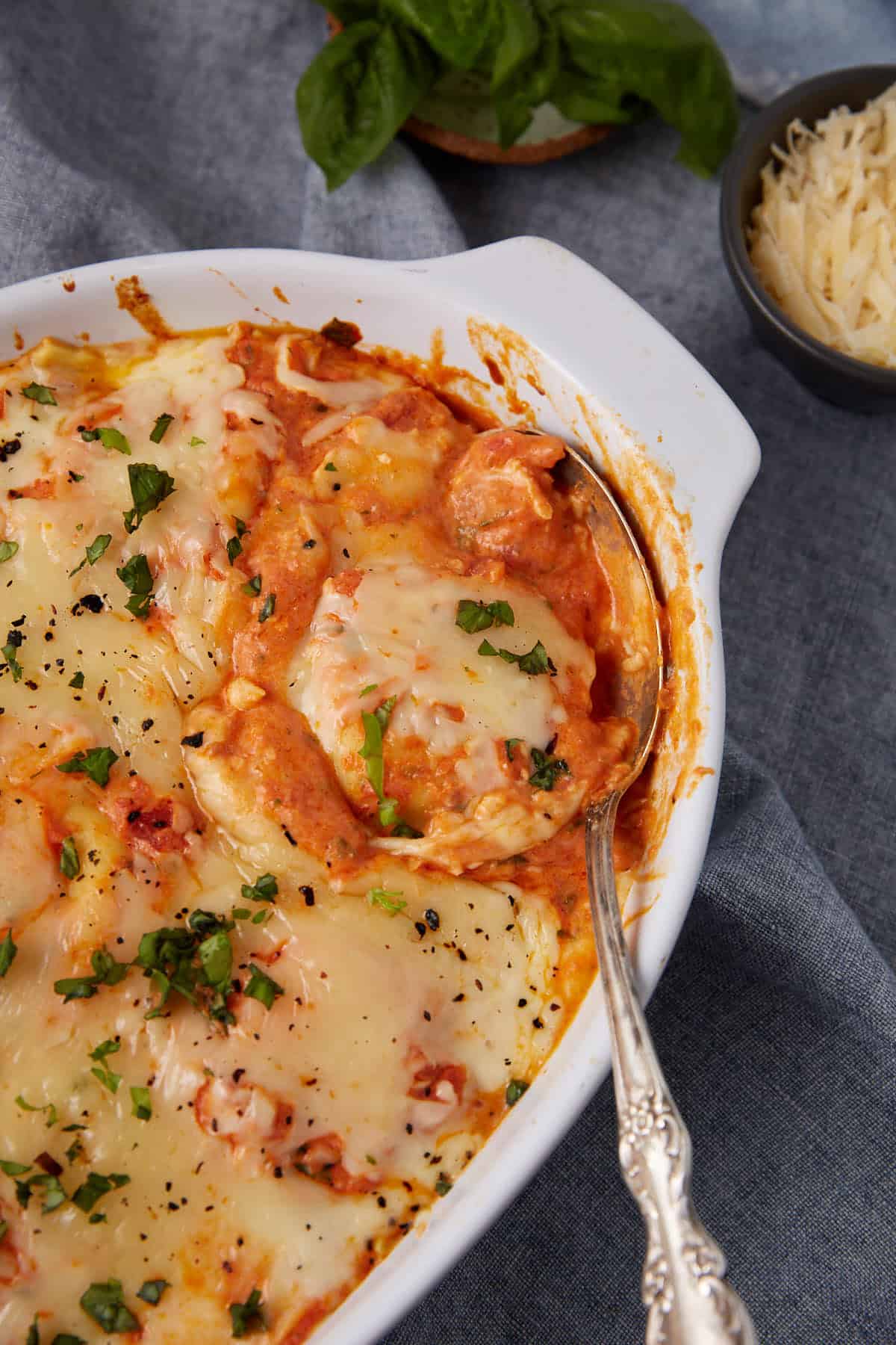 A baked dish of cheesy, saucy lasagna or Baked Ravioli with melted cheese and herbs on top, served in a white casserole dish with a silver serving spoon, next to a bowl of grated cheese and fresh basil.