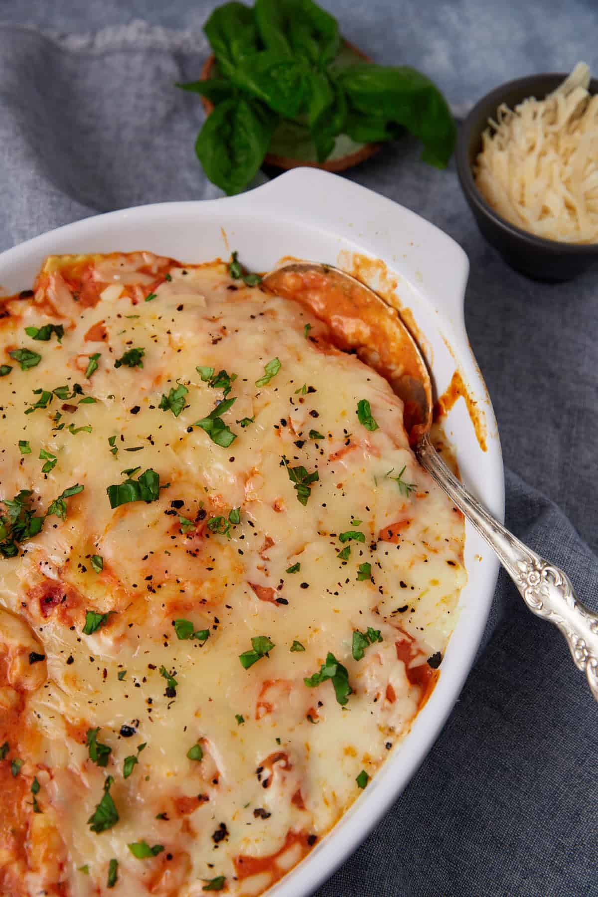 A baked dish of cheesy lasagna or Baked Ravioli, garnished with chopped parsley, with a serving spoon in the dish. In the background, a bowl of grated cheese and fresh basil leaves are visible.