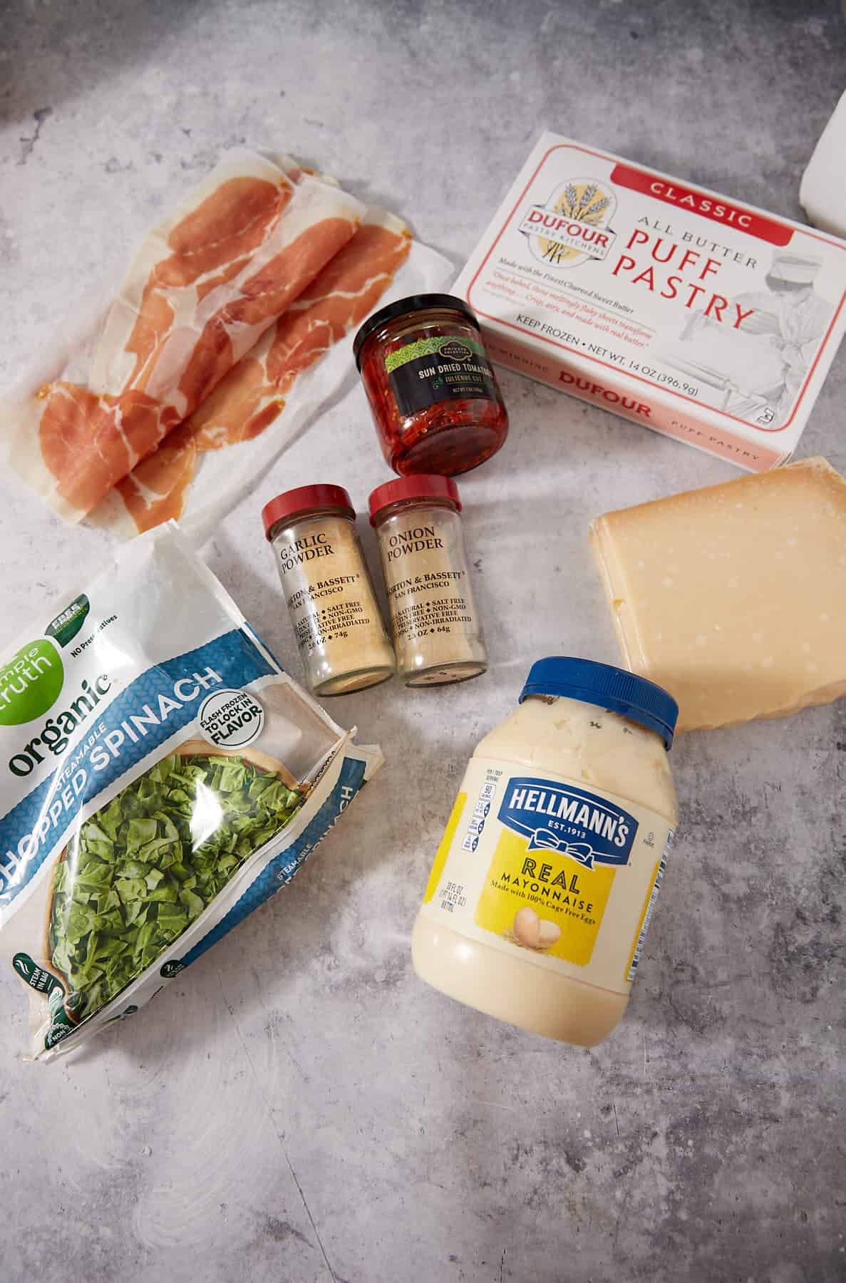 Ingredients for Puff Pastry Pinwheels are laid out on a gray surface: prosciutto, jarred red peppers, puff pastry, Parmesan cheese, mayonnaise, chopped spinach, garlic powder, and onion powder.