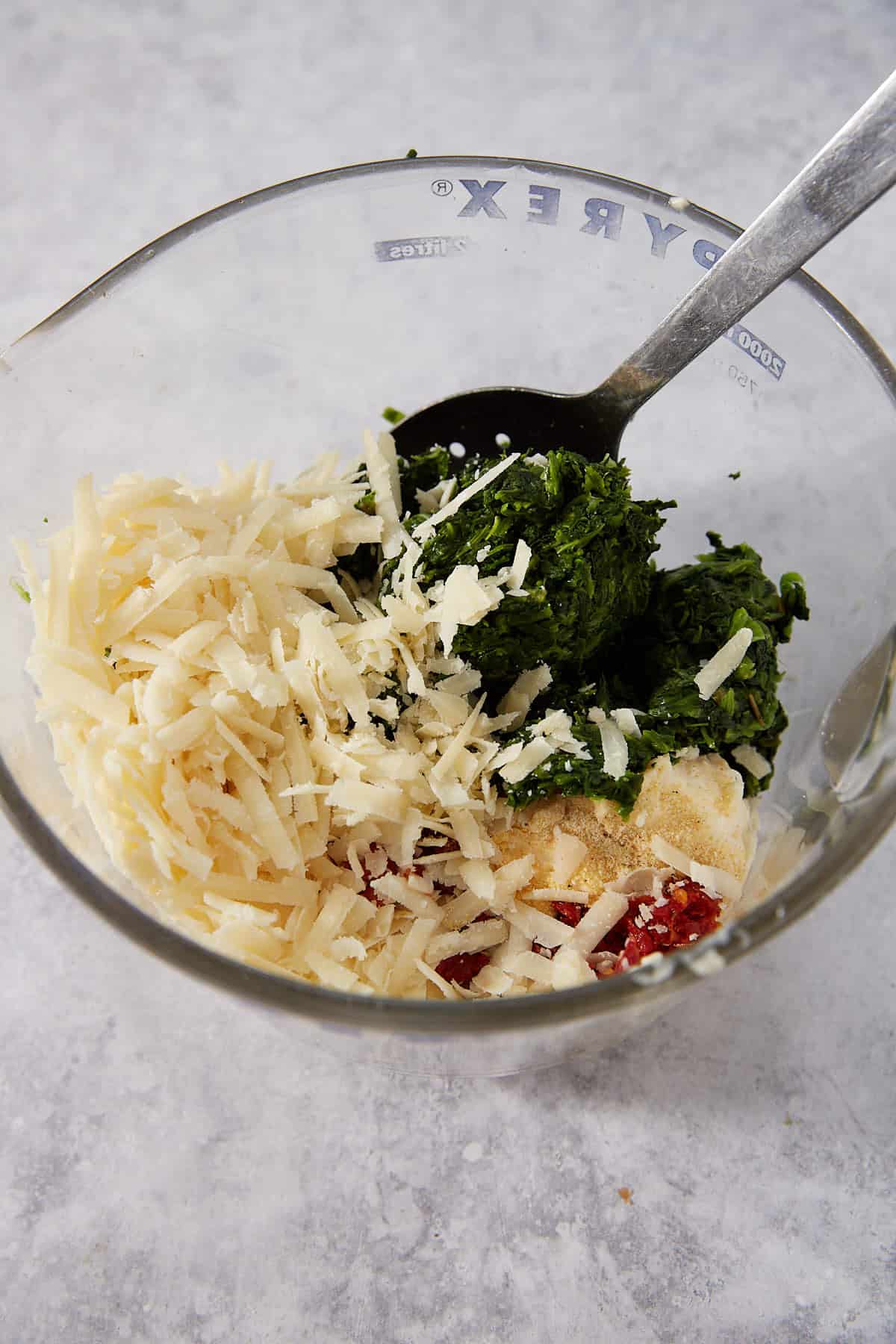 A glass bowl with shredded cheese, chopped spinach, red pepper flakes, and garlic powder—perfect for making Puff Pastry Pinwheels—sits on a light gray surface with a spoon inside.