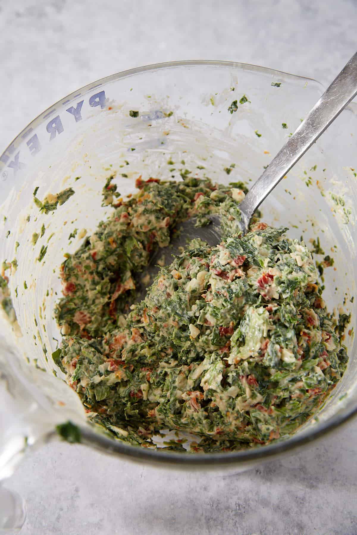 A glass mixing bowl filled with a creamy mixture of chopped leafy greens, bits of red peppers, and diced onions—perfect as a flavorful filling for Puff Pastry Pinwheels. A metal spoon rests inside the bowl.