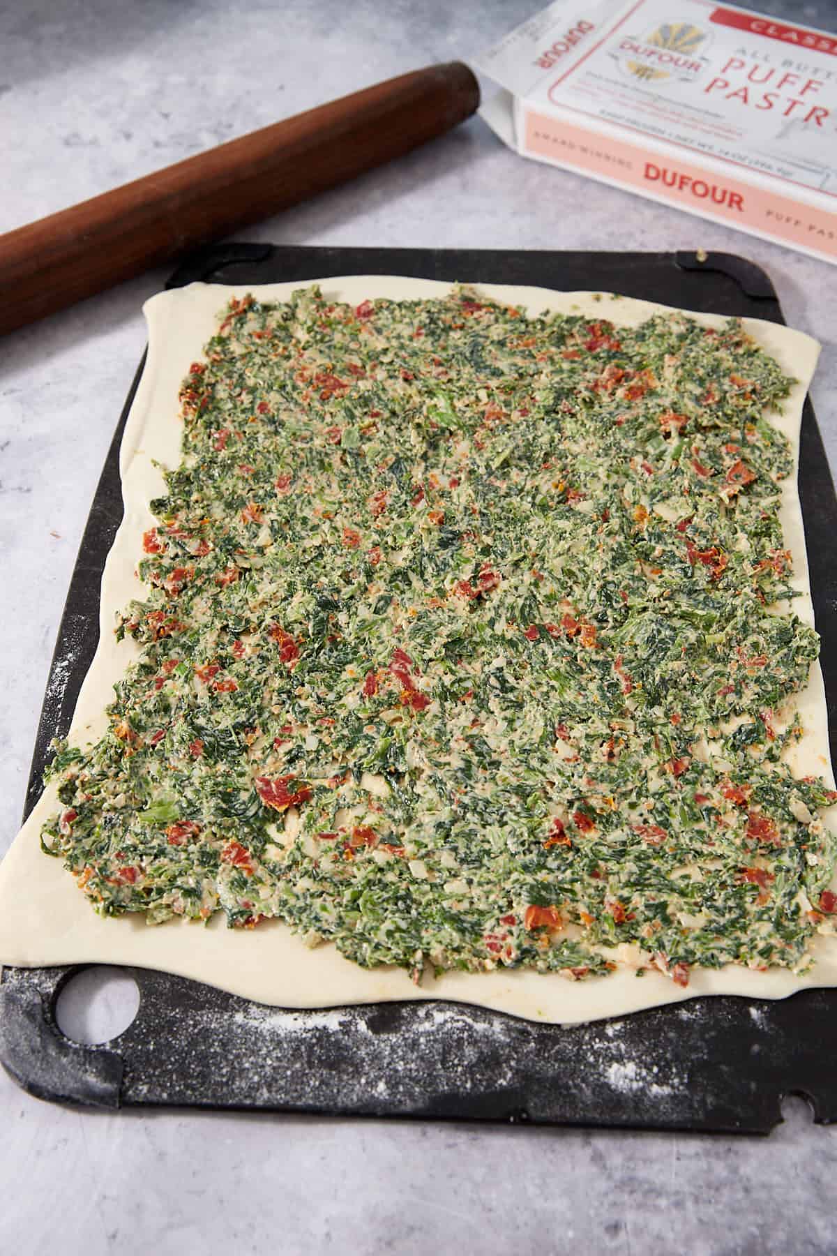 Rectangular puff pastry dough topped with a spread of finely chopped spinach, red peppers, and creamy filling is ready to be transformed into delicious Puff Pastry Pinwheels. Presented on a black cutting board, with a rolling pin and puff pastry box in the background, this setup showcases the perfect start for making these savory treats.