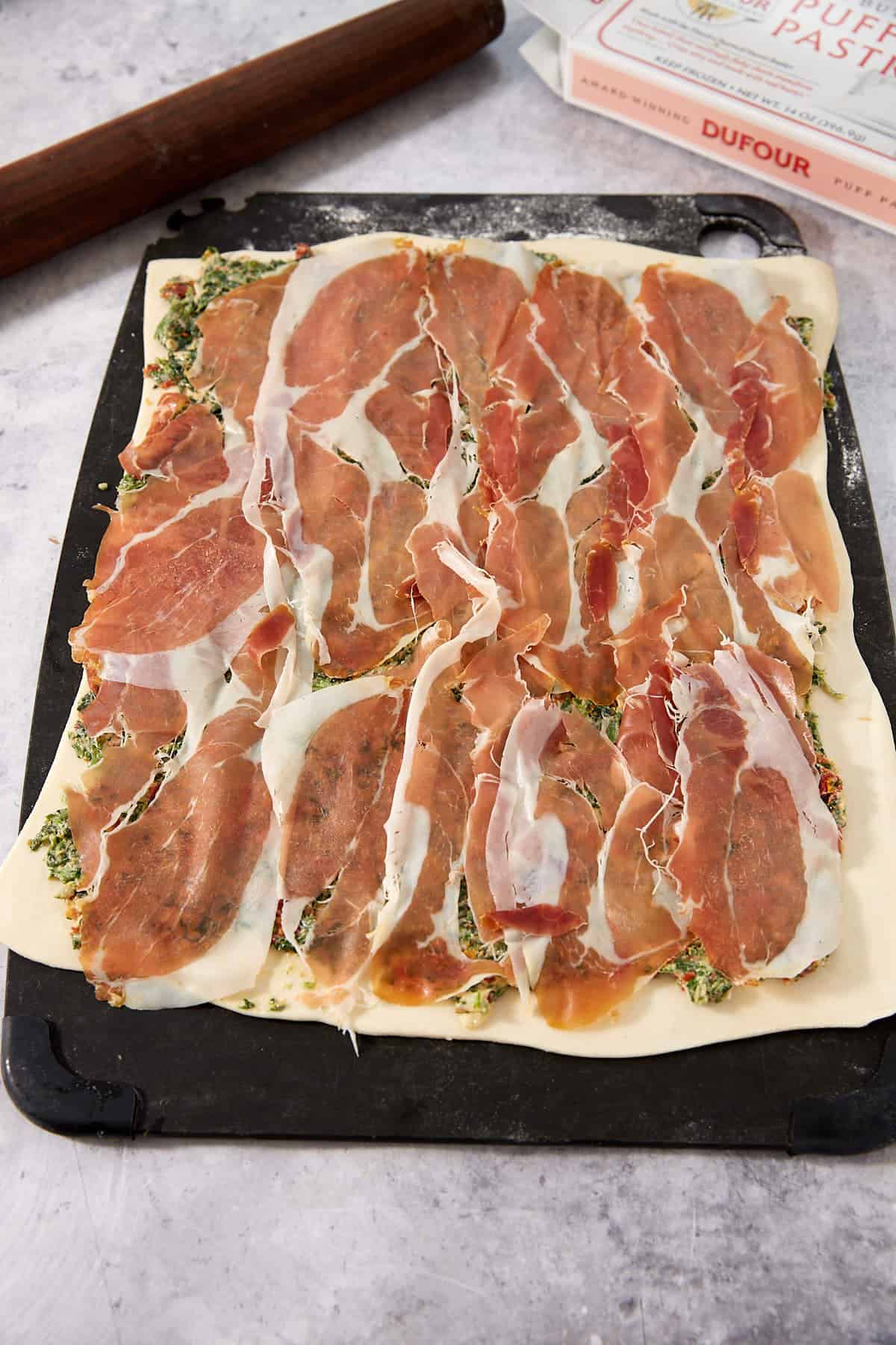 A sheet of puff pastry topped with a layer of thinly sliced prosciutto and green herb mixture, on a baking tray, is ready to be rolled into delicious Puff Pastry Pinwheels. A rolling pin and a pastry box are in the background.