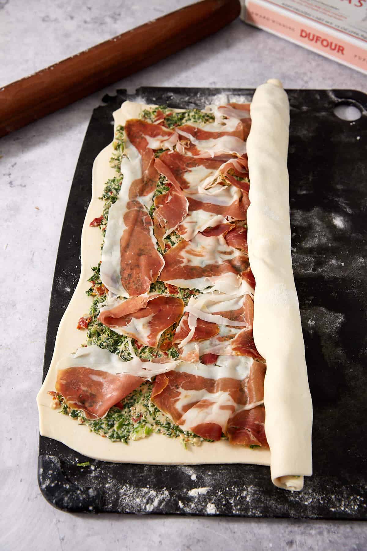 Rolled pastry dough filled with a spinach-herb mixture and slices of cured meat on a dark tray, partially rolled up to create delicious Puff Pastry Pinwheels, with a wooden rolling pin and a cookbook nearby on a light countertop.
