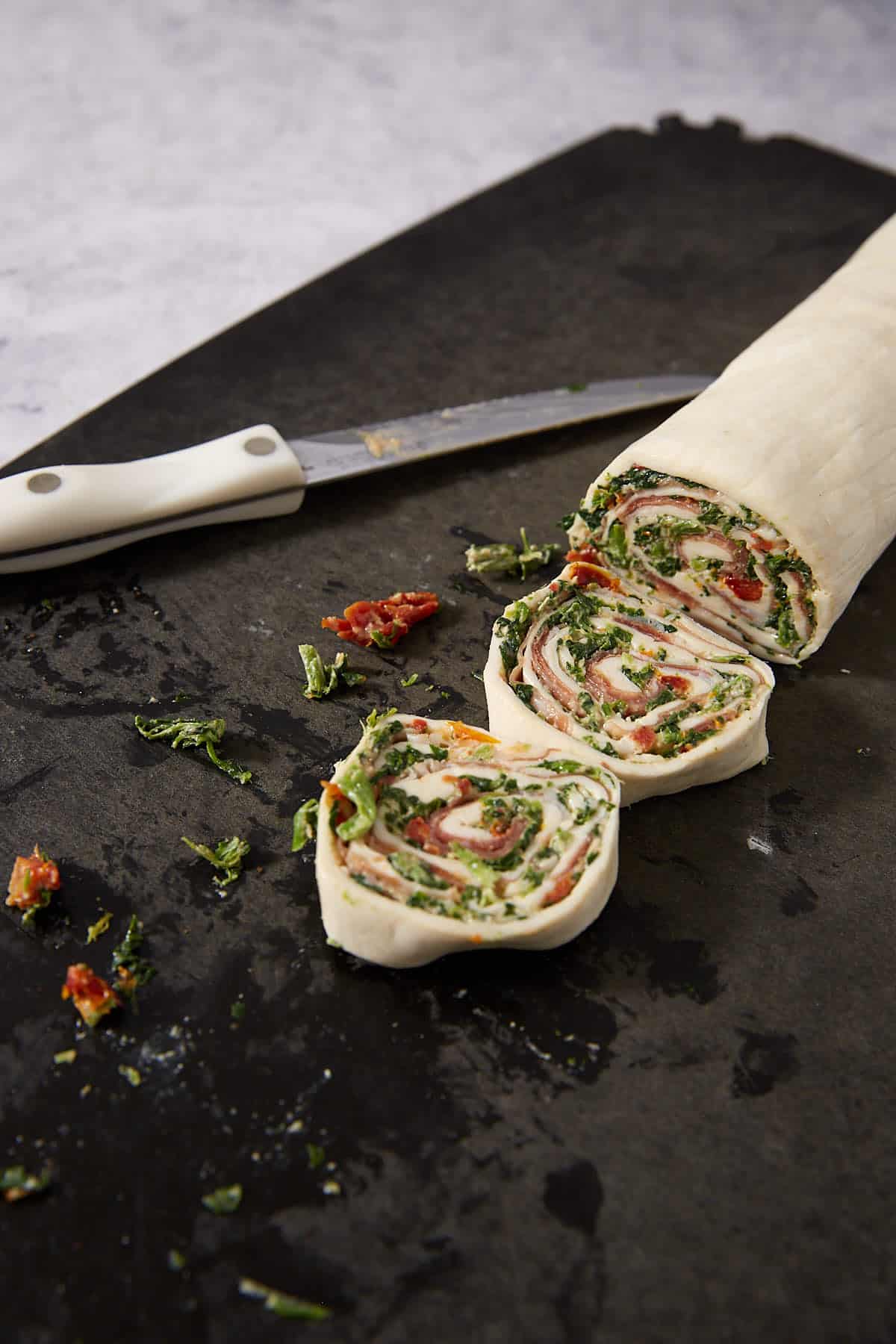 A rolled puff pastry pinwheels appetizer displays visible layers of spinach, sundried tomatoes, and meat on a dark surface, placed beside a white-handled knife.