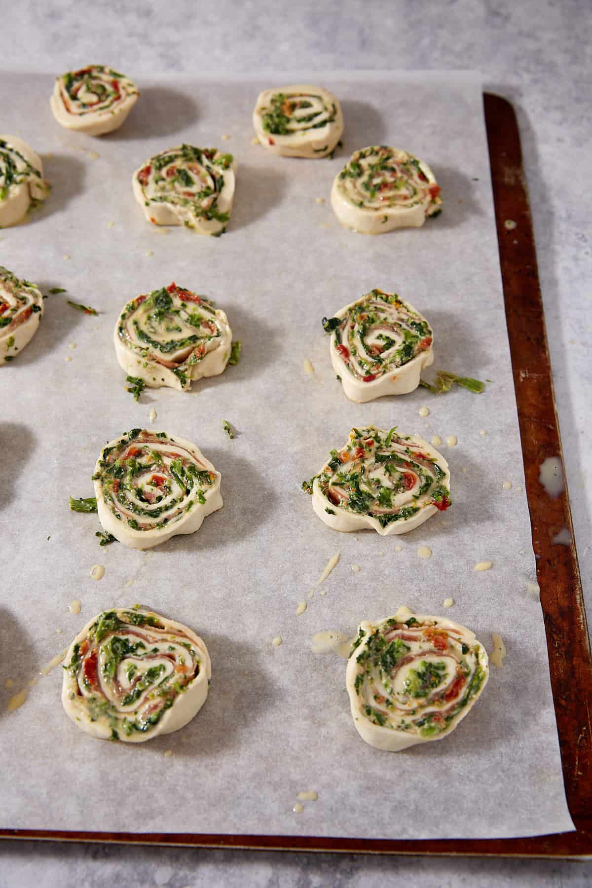 Unbaked Puff Pastry Pinwheels filled with green herbs and cheese are arranged on a parchment-lined baking sheet, ready to be baked.