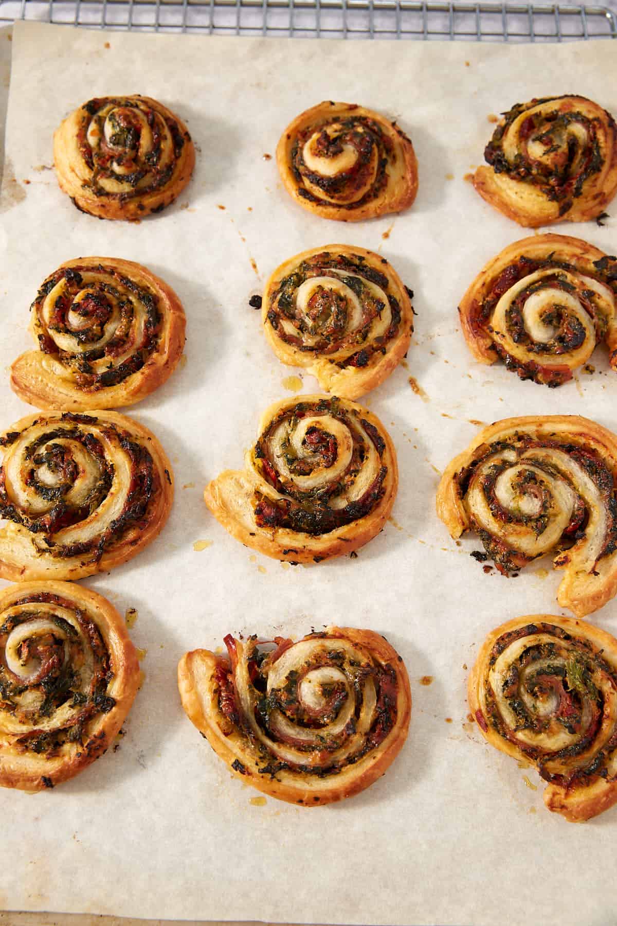 Twelve golden-brown Puff Pastry Pinwheels with herbs and cheese are arranged on parchment paper atop a cooling rack.