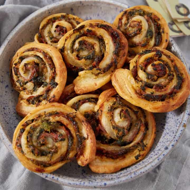 A speckled bowl filled with golden, spiral-shaped Puff Pastry Pinwheels containing savory herbs and greens, arranged on a light gray cloth.