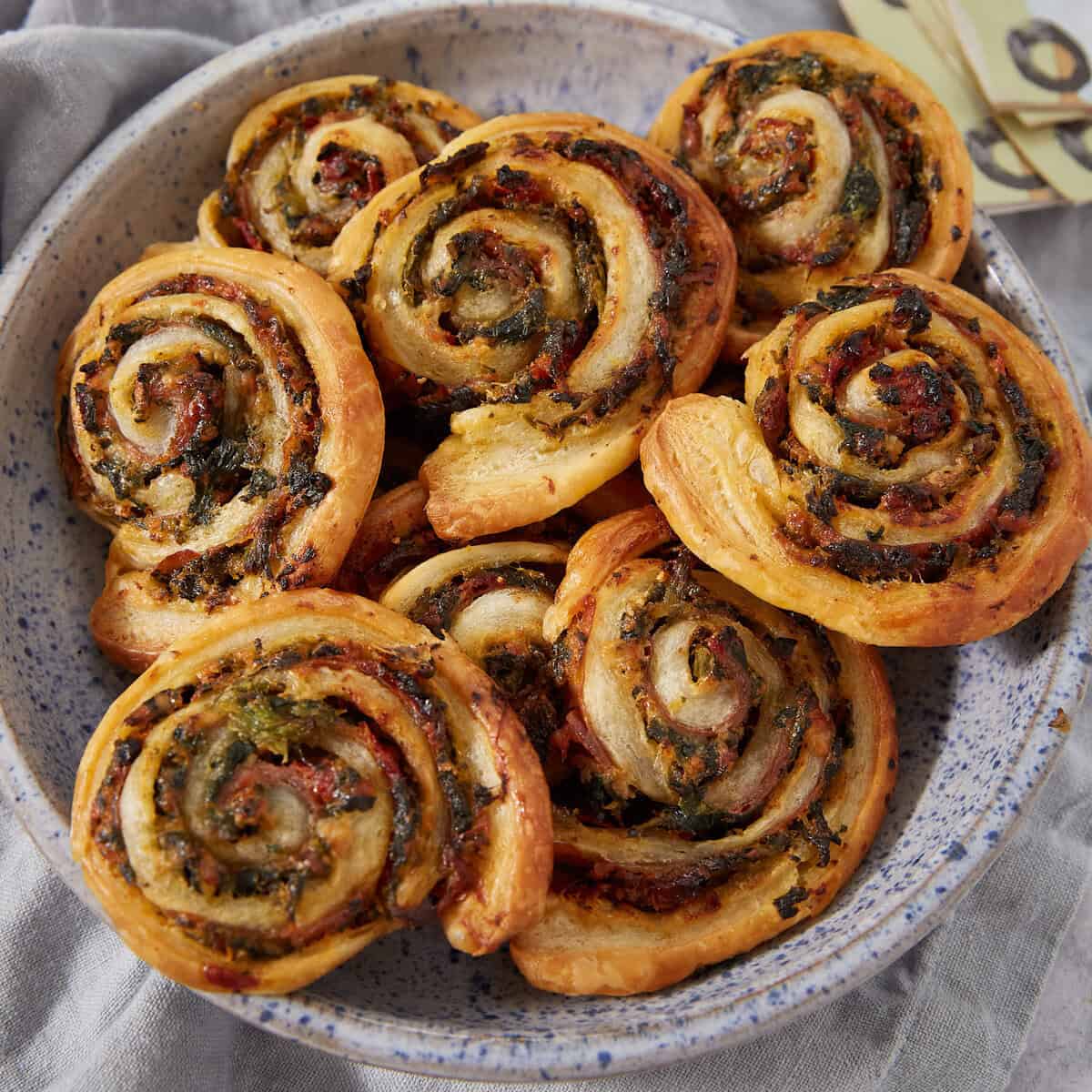 A speckled bowl filled with golden, spiral-shaped Puff Pastry Pinwheels containing savory herbs and greens, arranged on a light gray cloth.