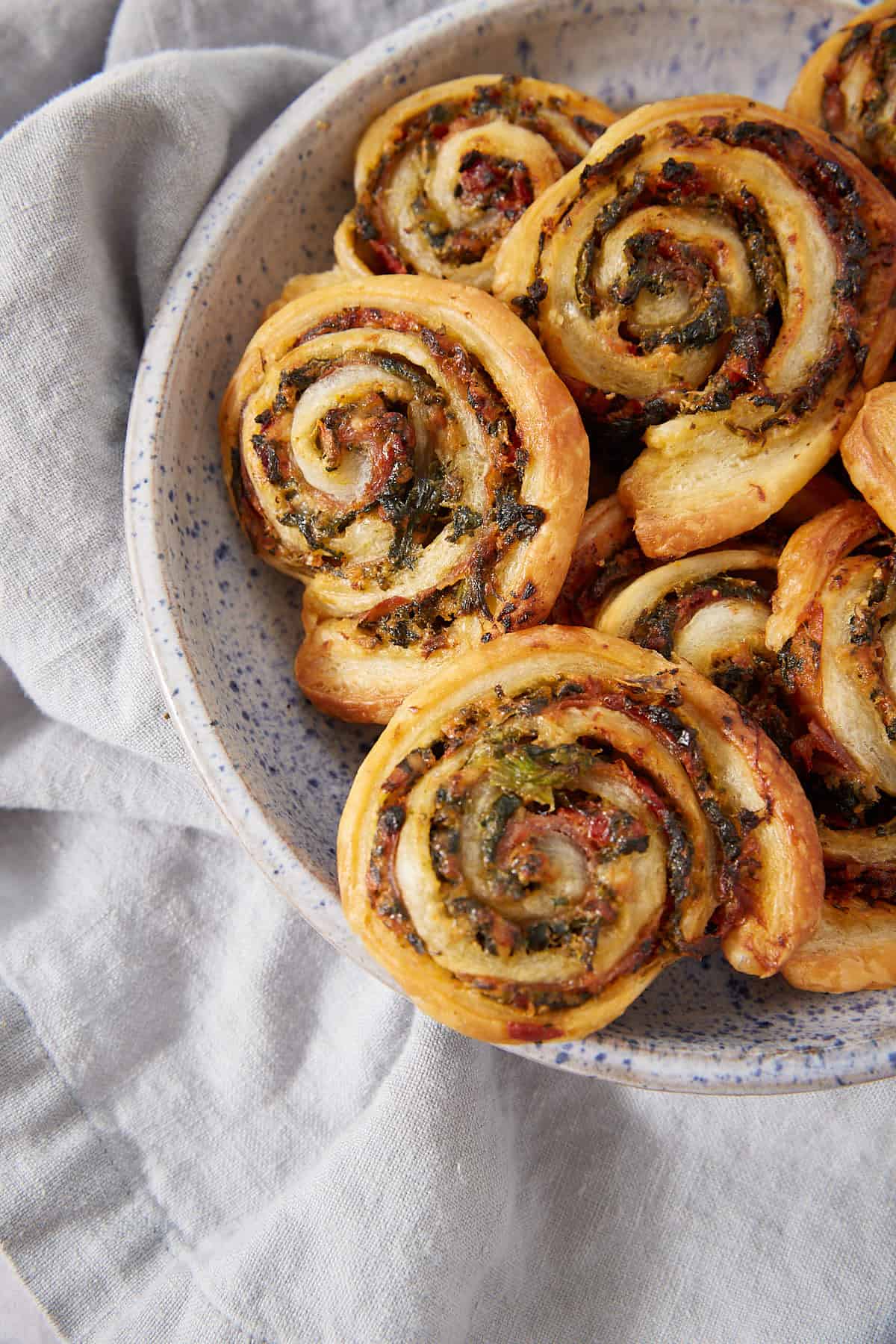 A speckled ceramic bowl filled with golden-brown Puff Pastry Pinwheels, their spiral shapes revealing vibrant green and red filling, sits on a light gray fabric surface.