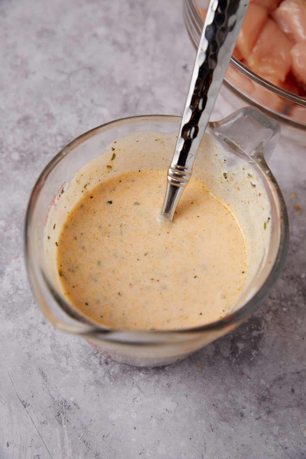 A glass measuring cup filled with creamy, seasoned marinade for Grilled Ranch Chicken with a metal spoon inside; a bowl of raw chicken pieces is partially visible in the background on a gray countertop.