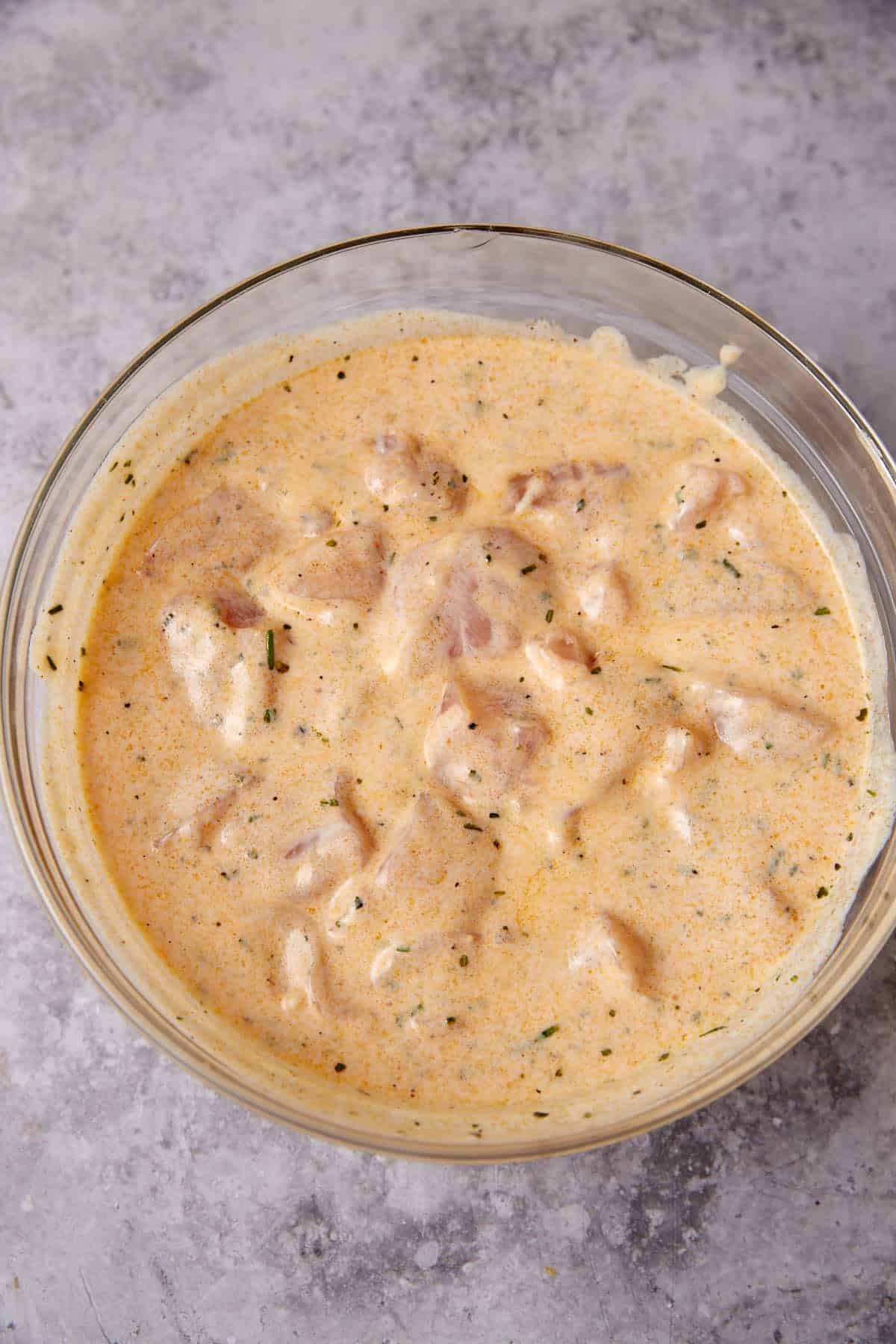 A glass bowl filled with pieces of raw chicken marinating in a creamy, orange-colored Grilled Ranch Chicken sauce with visible herbs, placed on a speckled gray surface.