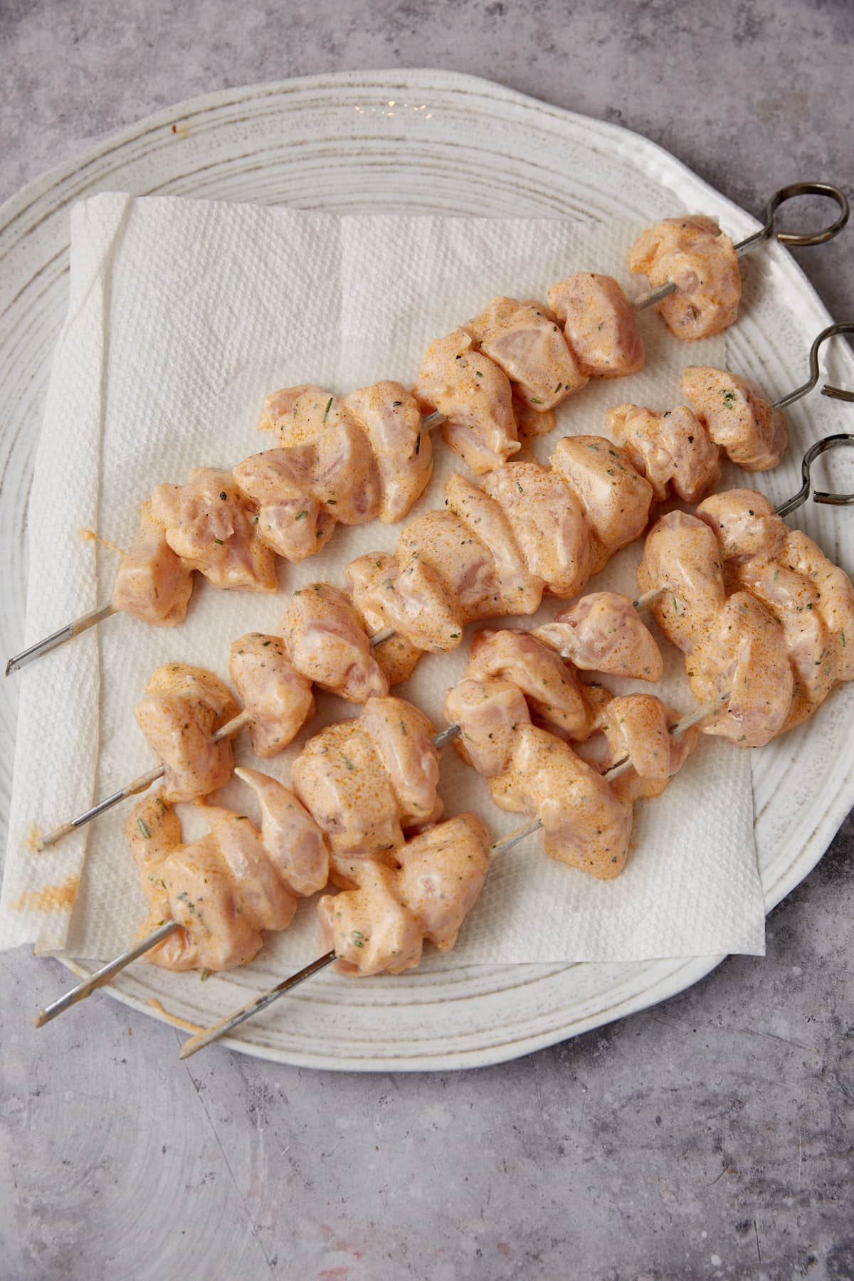 Four metal skewers with raw, marinated Grilled Ranch Chicken pieces are arranged on a white plate lined with paper towels, placed on a light gray surface.