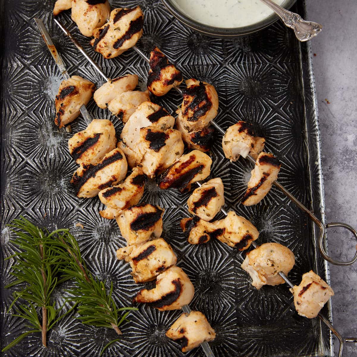 Grilled Ranch Chicken skewers with char marks are arranged on a patterned metal tray beside a sprig of fresh rosemary and a small bowl of creamy dipping sauce with a spoon.