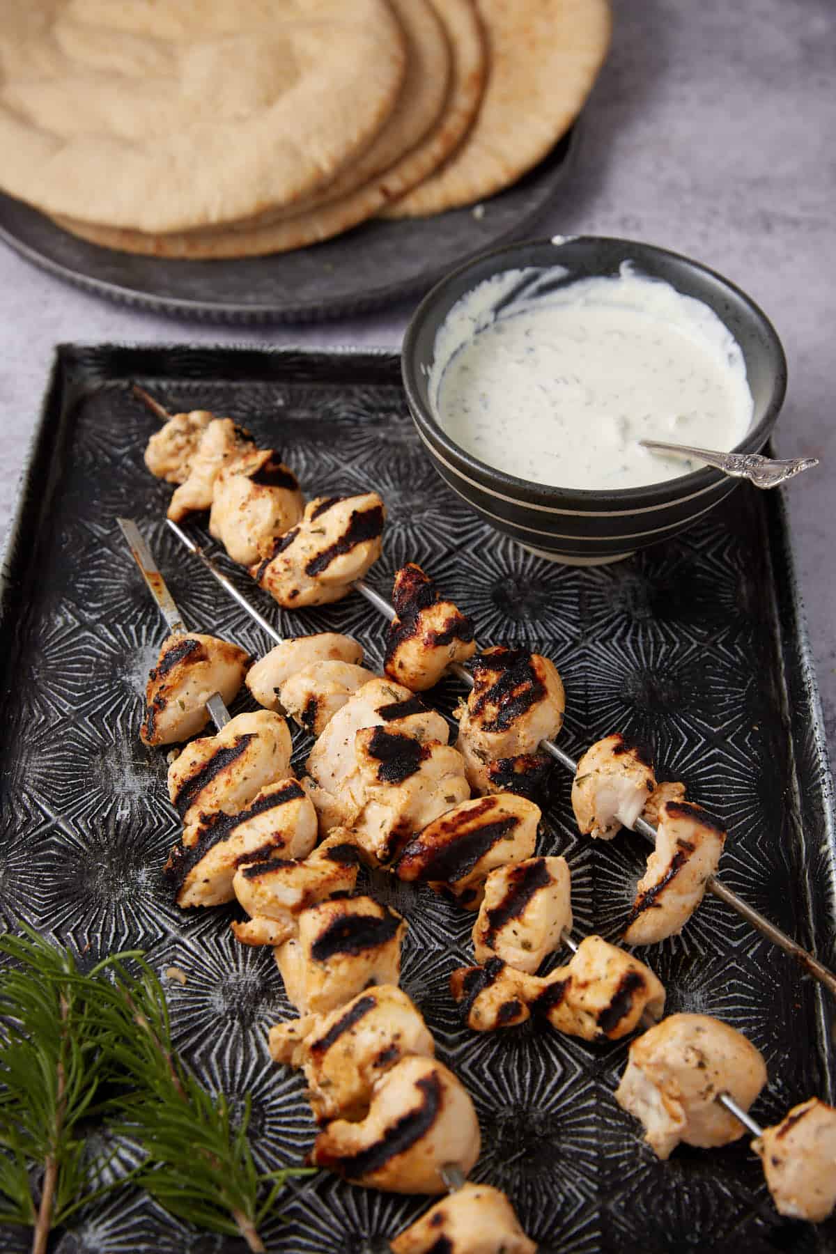 Grilled Ranch Chicken skewers on a patterned tray, served with a bowl of white dipping sauce and a stack of pita bread in the background. Fresh dill sprigs are placed beside the skewers.