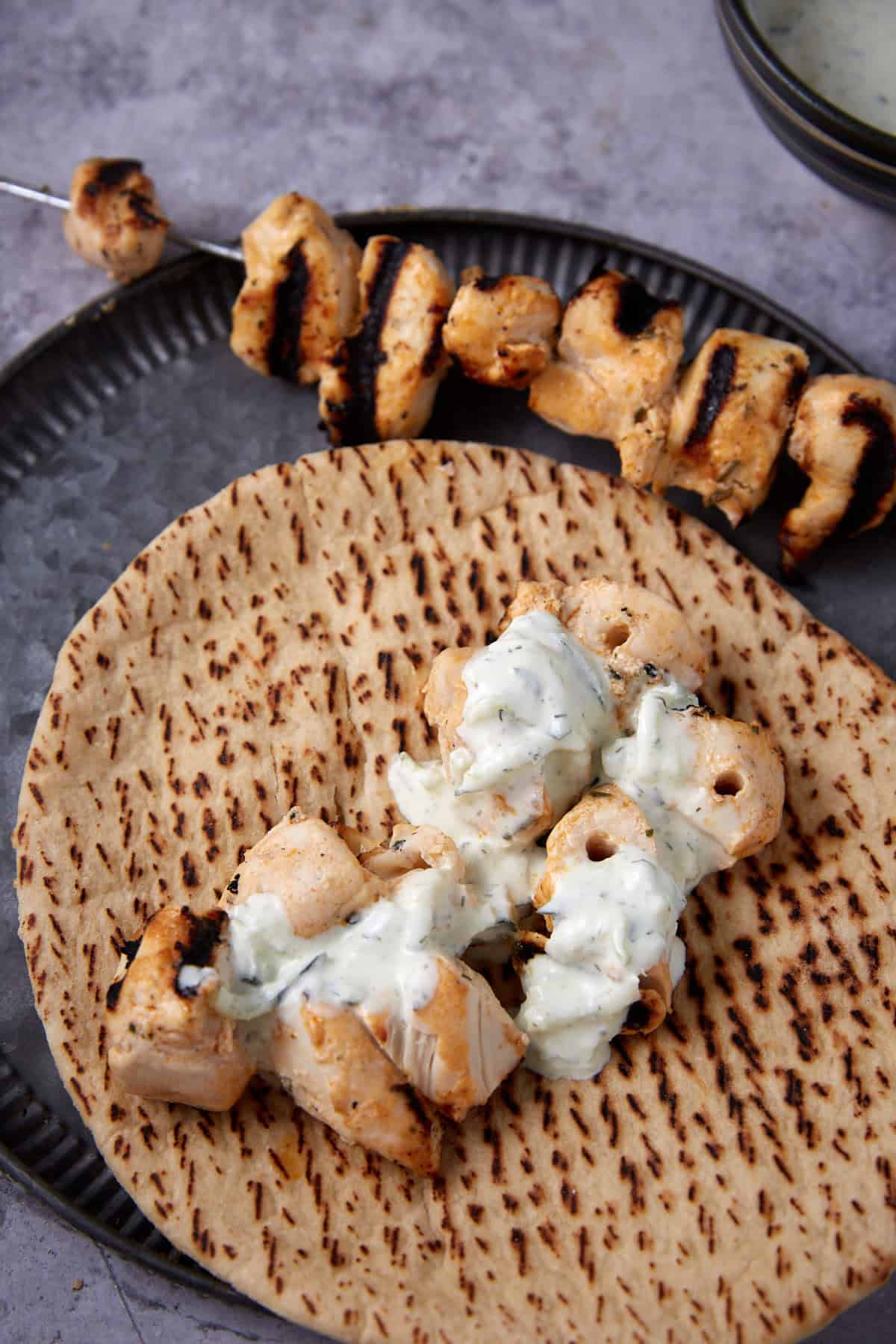 A grilled skewer of Grilled Ranch Chicken is placed on flatbread, topped with a generous dollop of creamy white sauce, with another chicken skewer seen in the background on a metal plate.