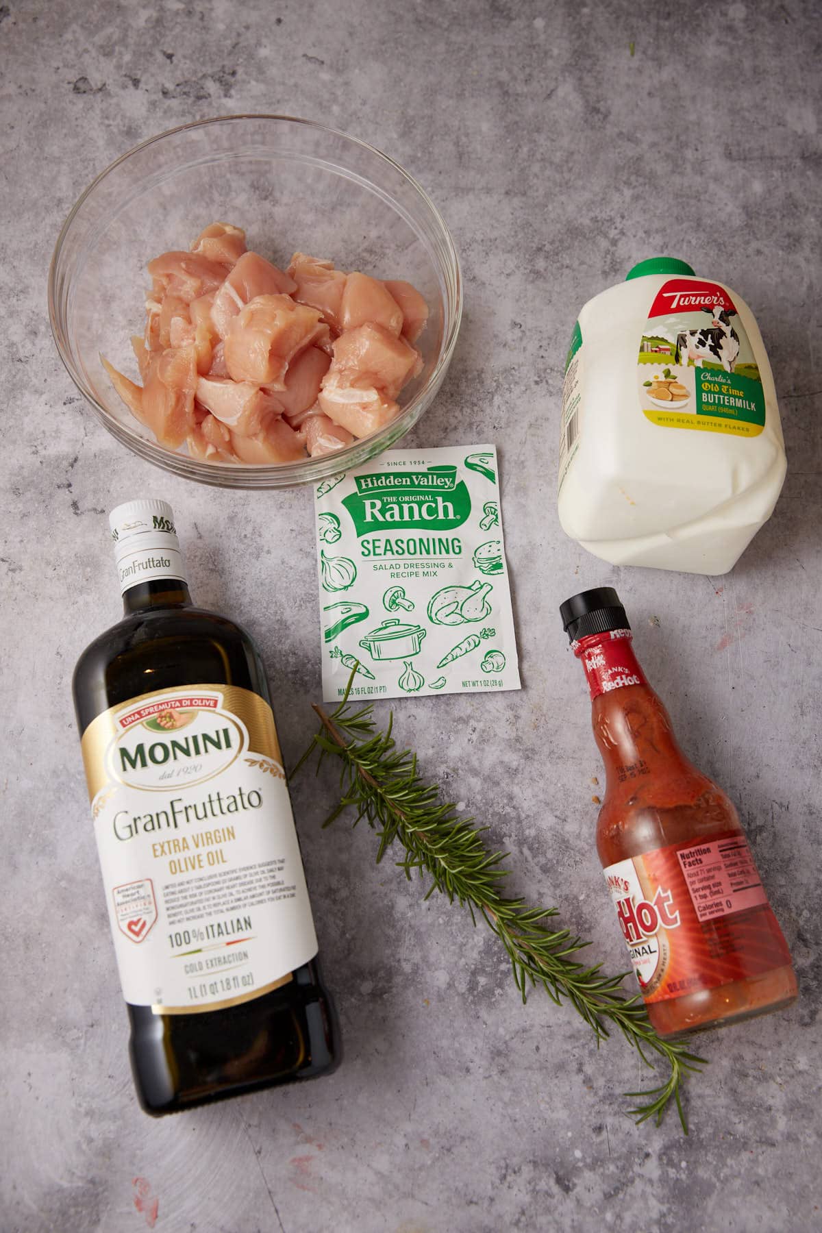 A bowl of raw cubed chicken for Grilled Ranch Chicken, a bottle of olive oil, ranch seasoning, milk, hot sauce, and a sprig of rosemary are arranged on a gray countertop.