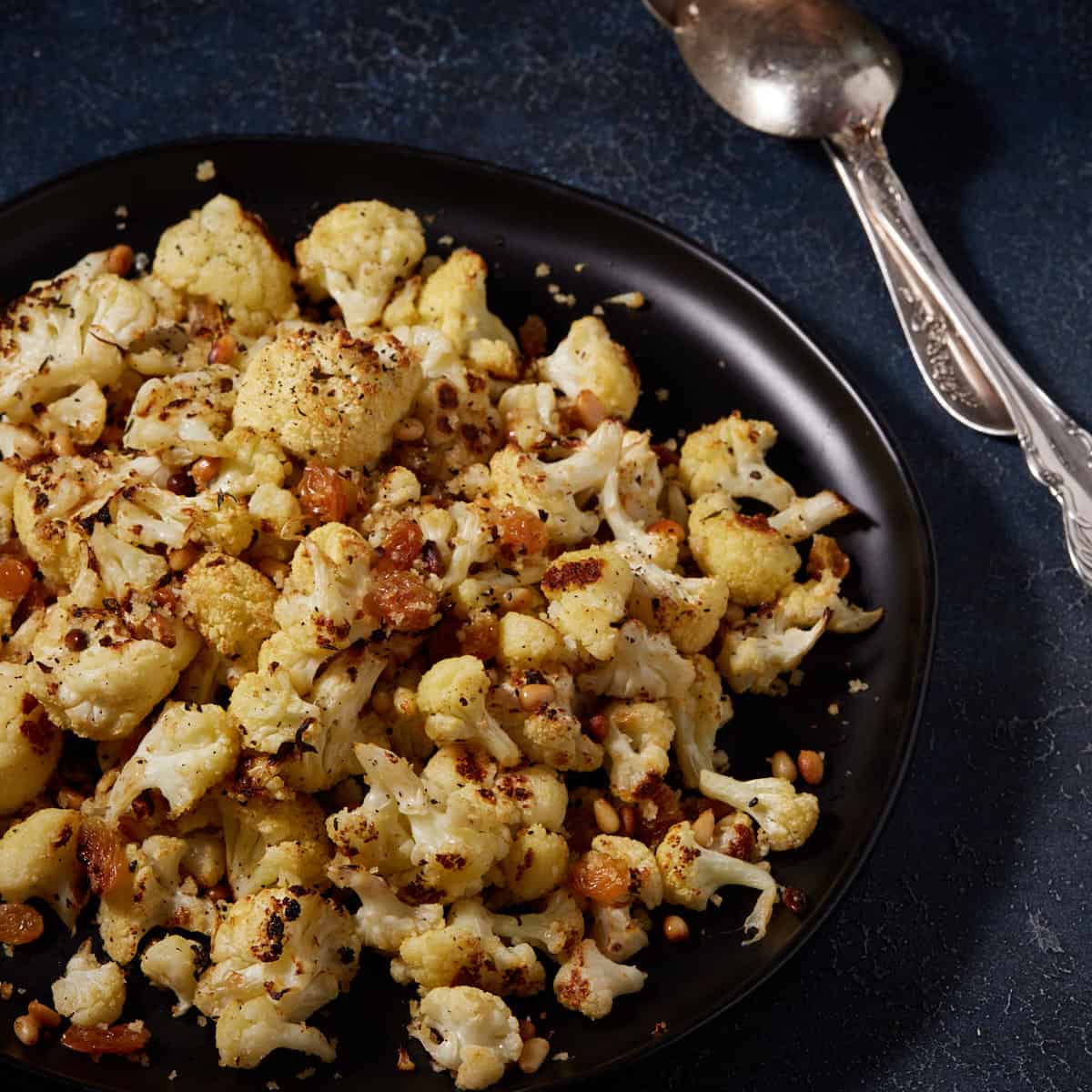 A black platter of roasted cauliflower with garlic next to serving spoons.