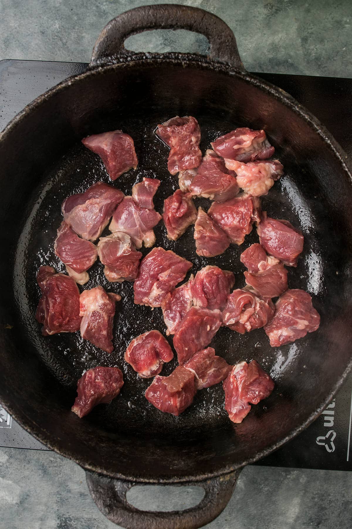 Raw chunks of red meat, perfect for Beef Bourguignon, are spread out in a black cast iron skillet, starting to brown as they cook. The skillet sits on a stovetop with a gray countertop in the background.