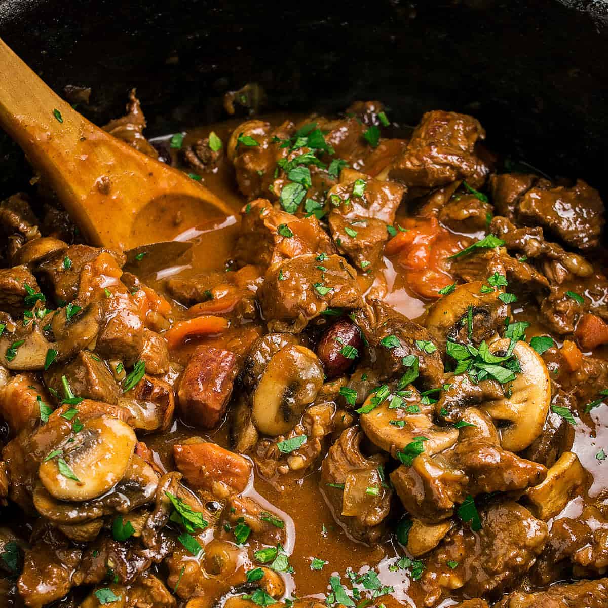 A close-up of a hearty Beef Bourguignon stew with mushrooms, carrots, and chunks of beef in a rich brown sauce, garnished with chopped parsley. A wooden spoon is stirring the savory mixture inside a pot.