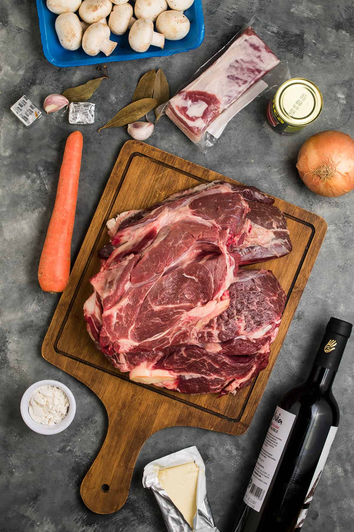 A wooden cutting board with raw beef steaks for Beef Bourguignon, surrounded by a carrot, mushrooms, onion, bay leaves, garlic, wine, butter, flour, broth cubes, tomato paste, and a pack of bacon on a gray surface.