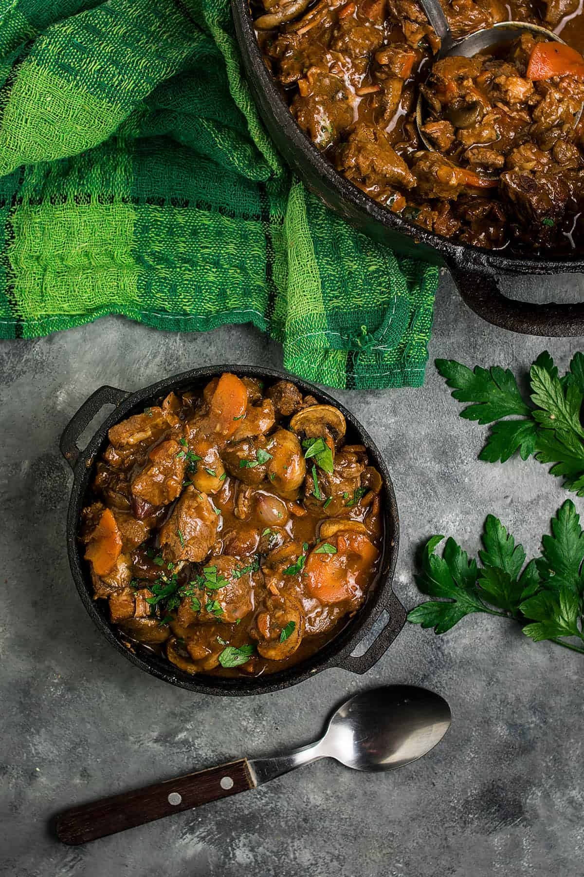 A bowl of hearty Beef Bourguignon with carrots and herbs sits on a gray surface near a spoon, a green towel, and fresh parsley, with a pot of more stew partially visible above.