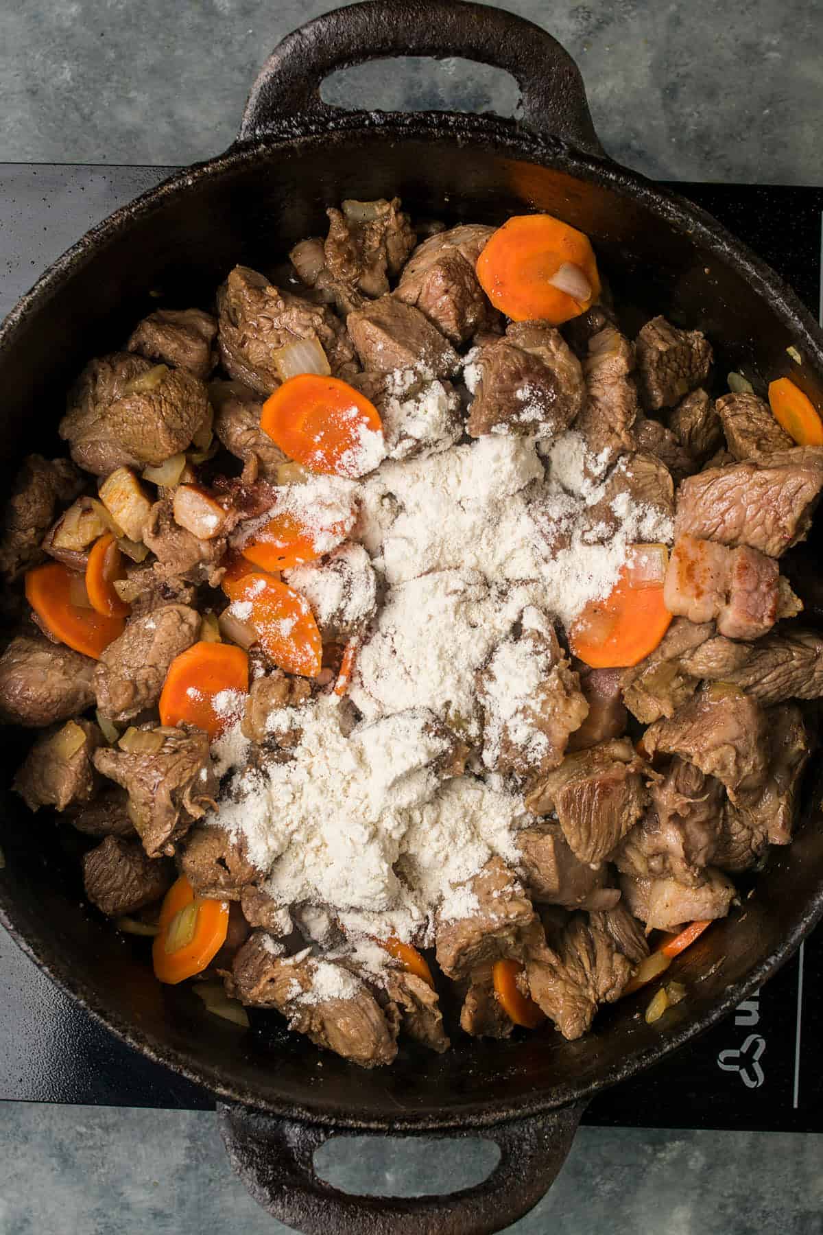 Chunks of browned beef, sliced carrots, and chopped onions simmer in a cast-iron pot for Beef Bourguignon, with a mound of white flour sprinkled on top, ready to be mixed.