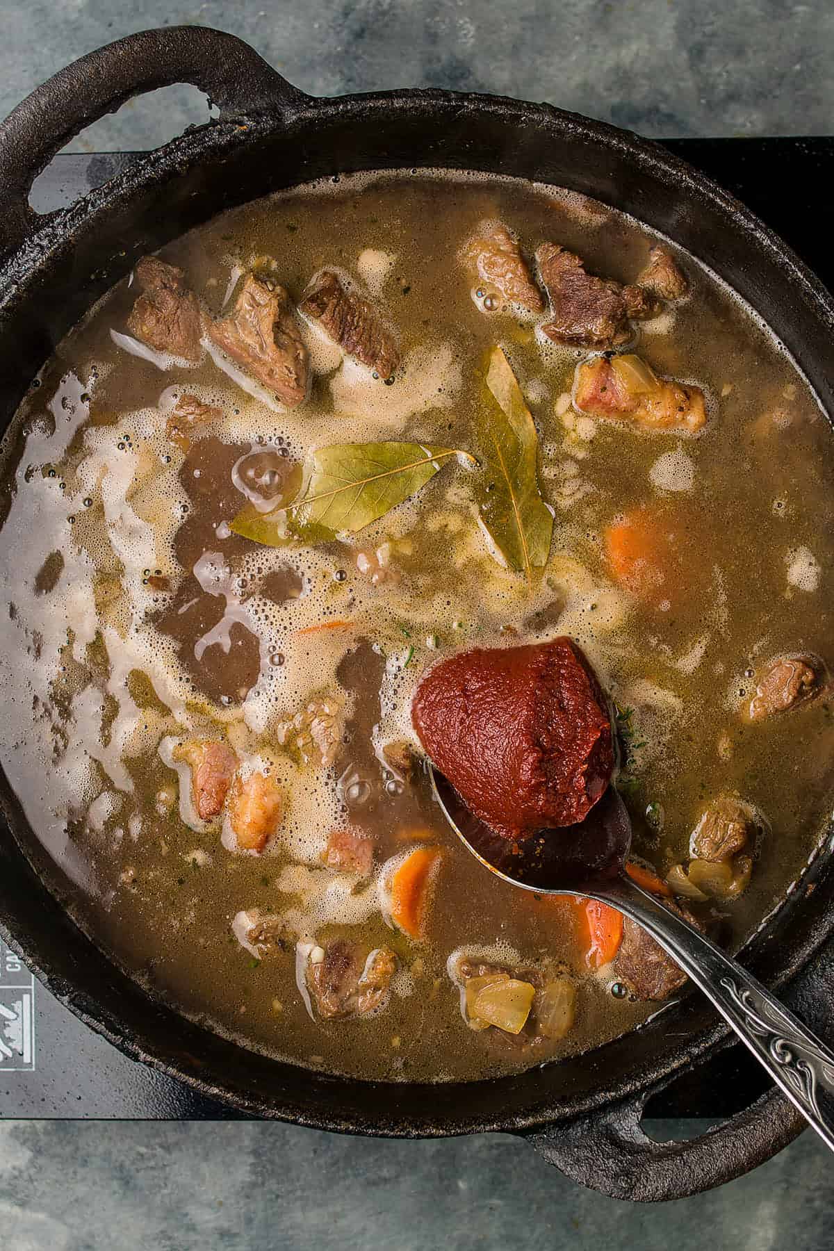 A pot of Beef Bourguignon with chunks of meat, carrots, and bay leaves simmers on the stove. A spoon holding a dollop of tomato paste is being added to the stew.