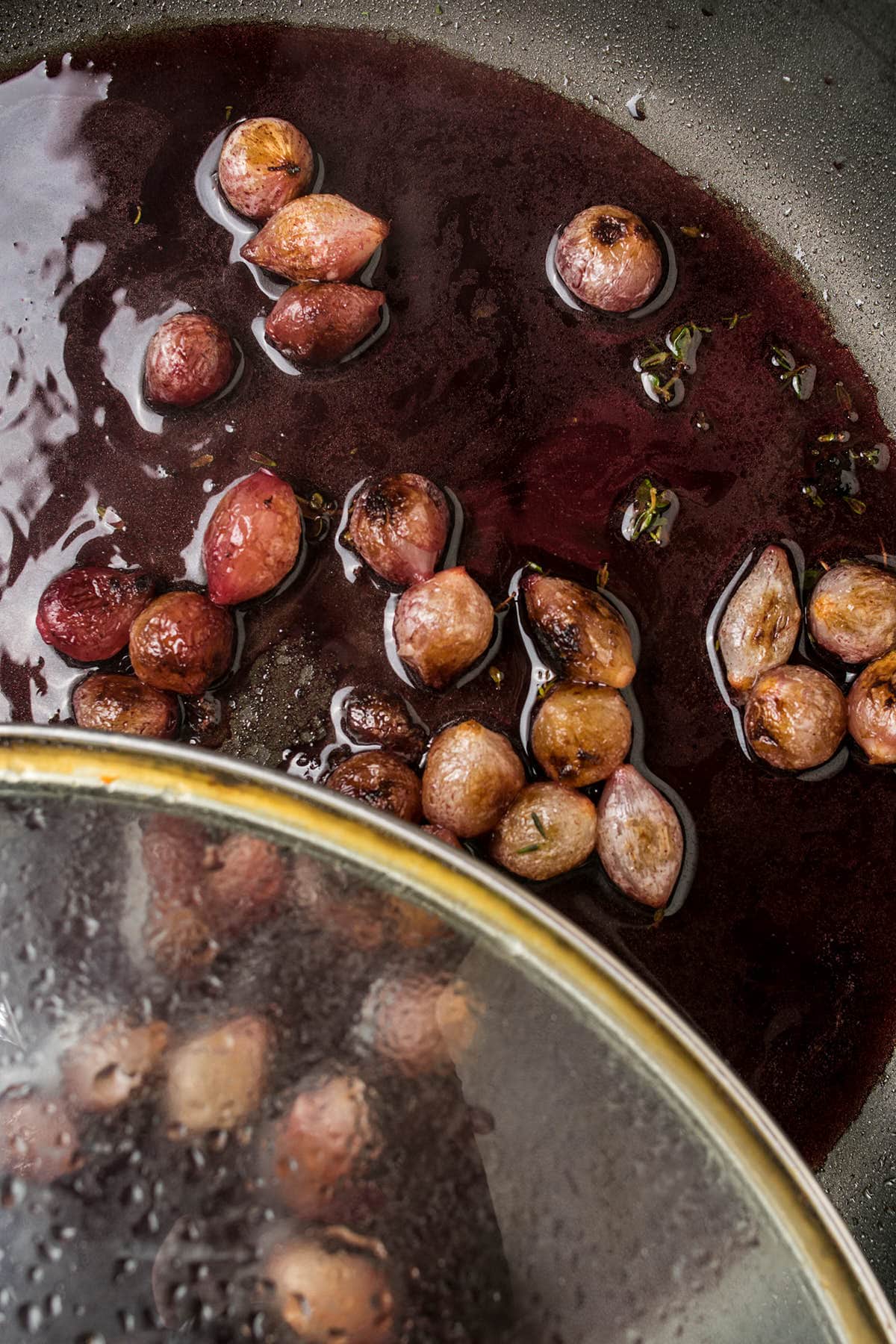 Red grapes are simmering in a dark, glossy liquid inside a pan. A glass lid, partially covering the pan, shows steam and moisture—evoking the rich aroma of Beef Bourguignon as the grapes gently poach in a luscious sauce.