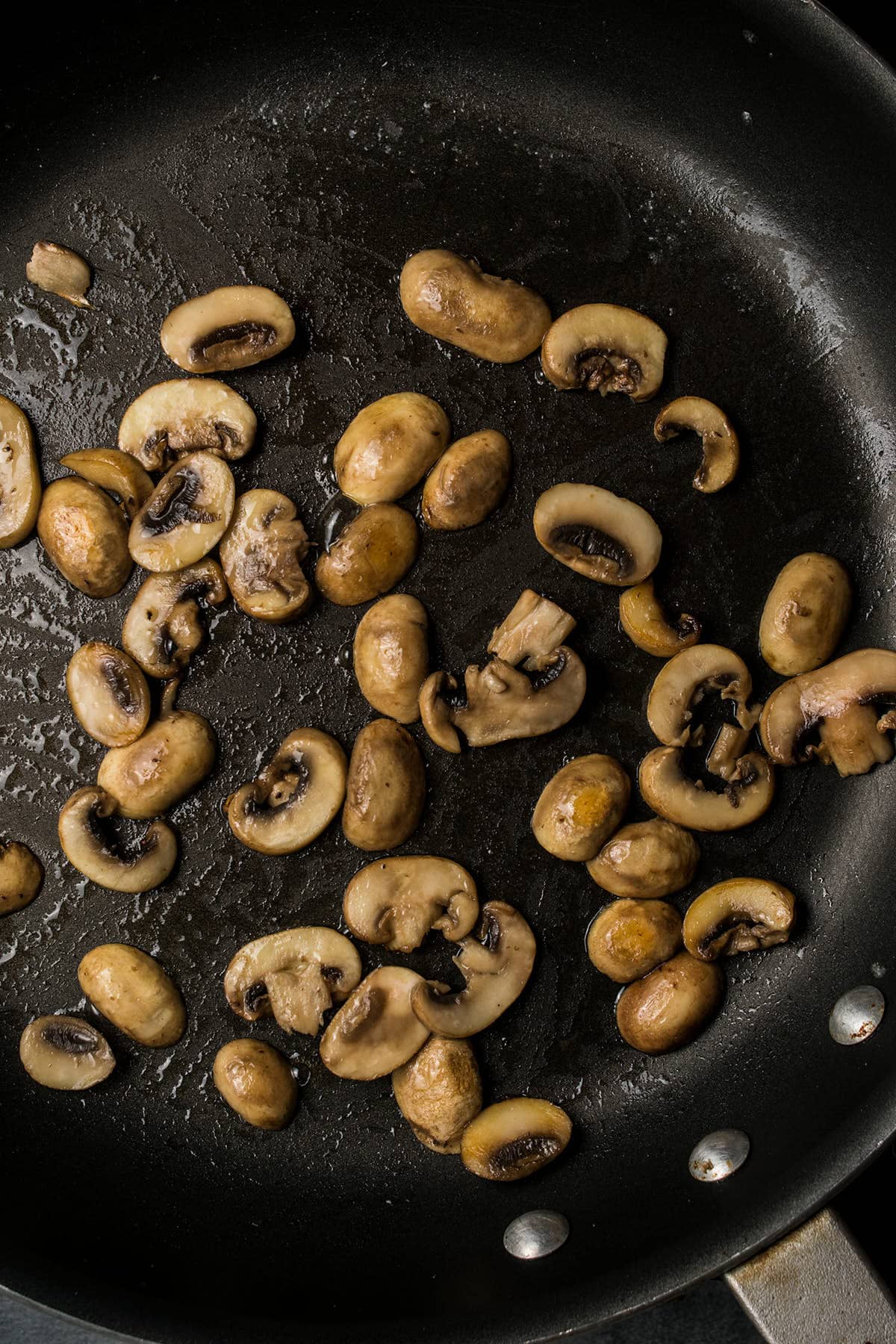 Sliced and whole mushrooms sautéing in a black skillet with some oil, showing light browning and a few scattered pieces—perfect for adding richness to a classic Beef Bourguignon.