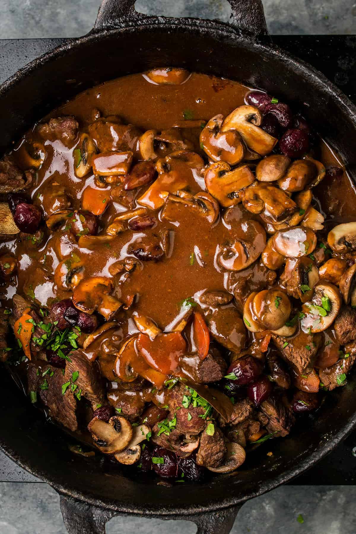 A cast iron pot filled with Beef Bourguignon, featuring a rich brown sauce, tender beef stew with sliced mushrooms, carrots, and a garnish of chopped parsley.